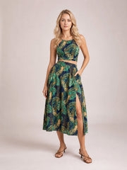 A model poses in a navy, green, and gold printed cami and slit skirt set with slender straps, a cropped fit, high waist, flared midi silhouette, and side slit.