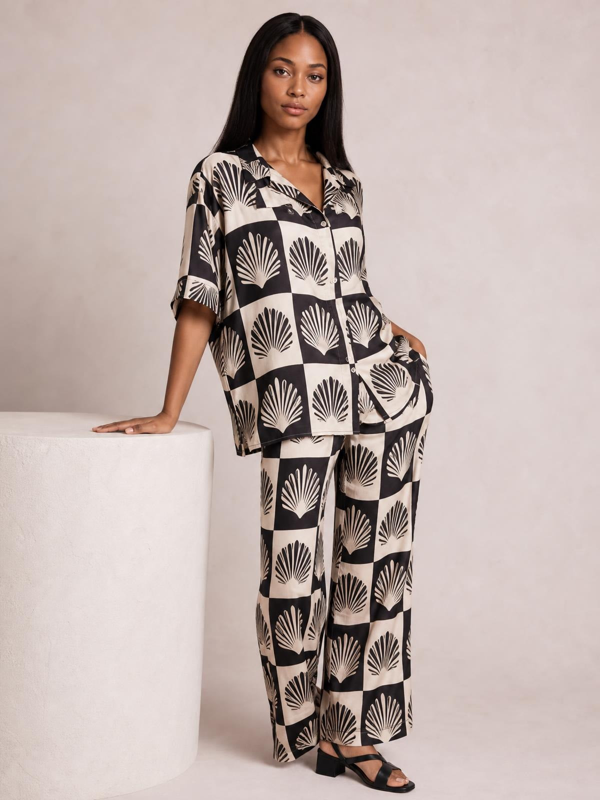 Model poses in a black printed collared top with button front and matching flowing wide-leg pants, leaning one hand on a pedestal in a coordinated set.