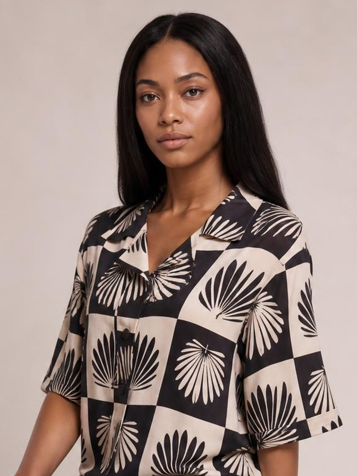 Model wearing a black printed collared neck top with bold floral motifs, button front, short sleeves, and a relaxed silhouette, facing the camera.