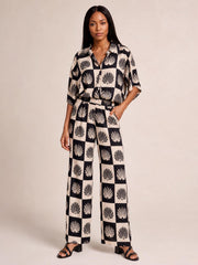 Model poses with one hand in a pocket, wearing a black printed collared buttoned top and matching wide-leg pants with a flowing silhouette.