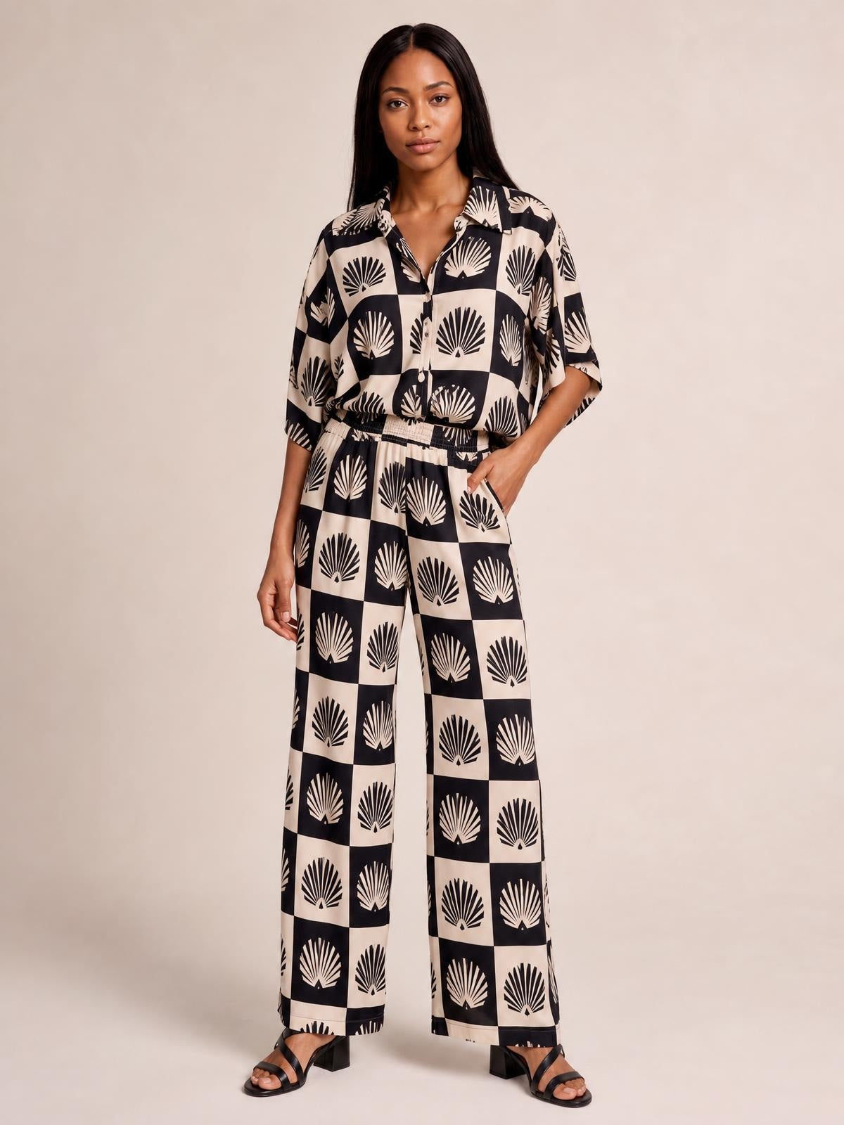 Model poses with one hand in a pocket, wearing a black printed collared buttoned top and matching wide-leg pants with a flowing silhouette.