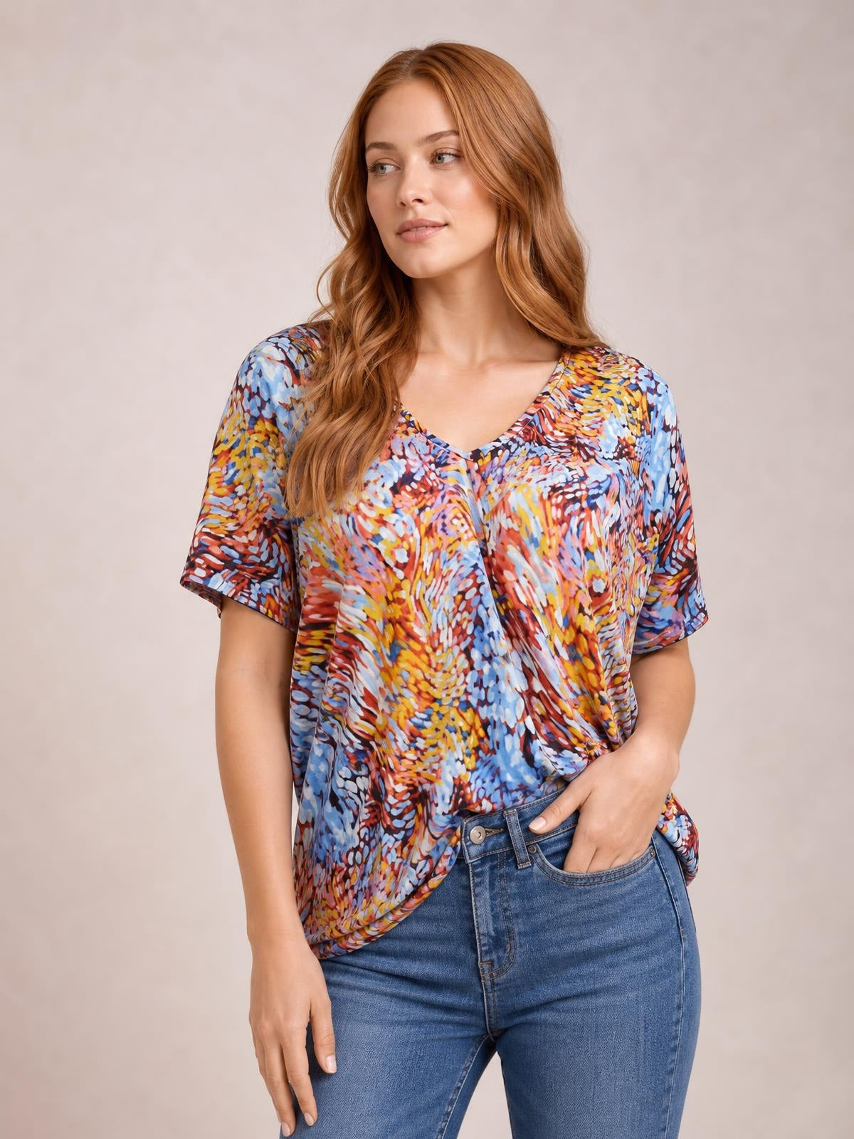 Model wears a multicolored printed dolman flowy top with a V-neck, relaxed draped silhouette, and short sleeves, posing with one hand in her jeans pocket.