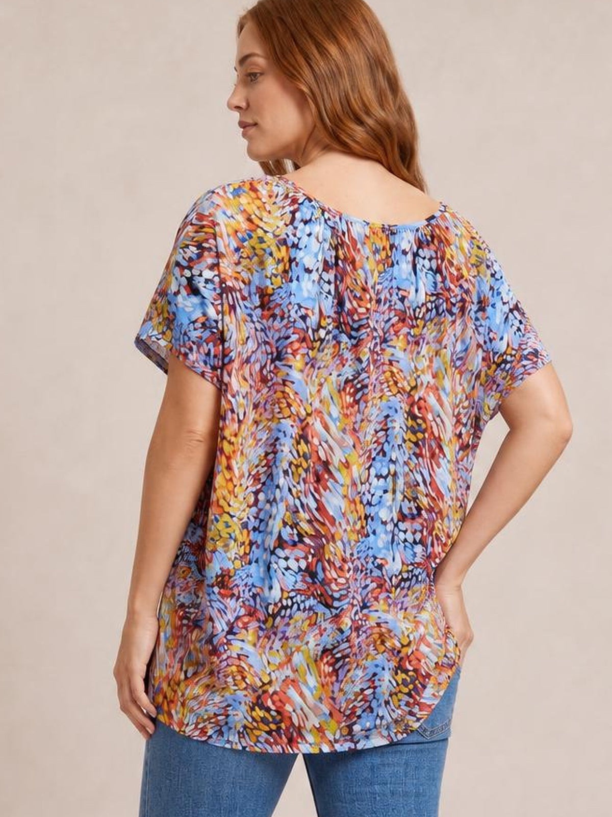 Model poses with one hand on her hip, showing the back of a long, relaxed dolman-sleeve top in a vibrant multicolored print.
