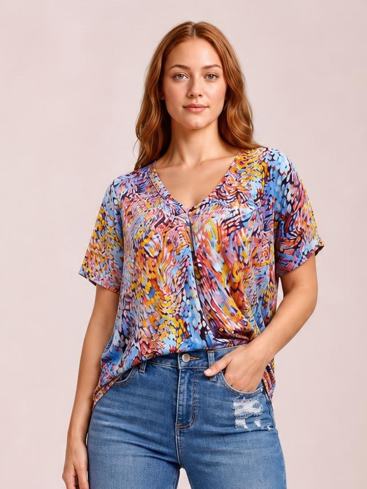 Model wearing a multicolored printed dolman flowy top with a V-neck, short sleeves, relaxed silhouette, and draped hem, posing with one hand in her jeans pocket.
