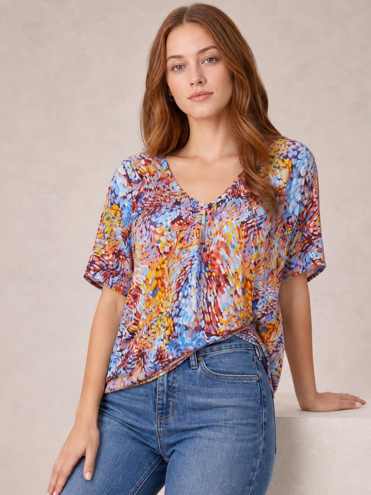 Model wearing a multicolored printed dolman flowy top with a V-neck, short sleeves, relaxed long silhouette, and draped hem.