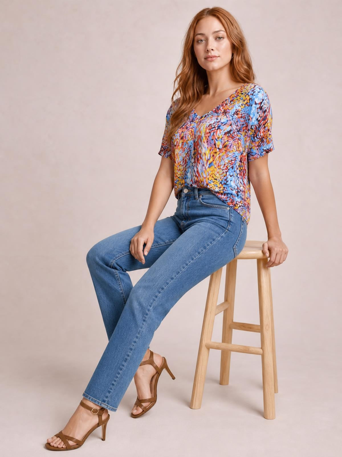 A seated model wears a multicolored printed dolman top with a V-neck, short sleeves, relaxed flowy silhouette, and softly draped hem.