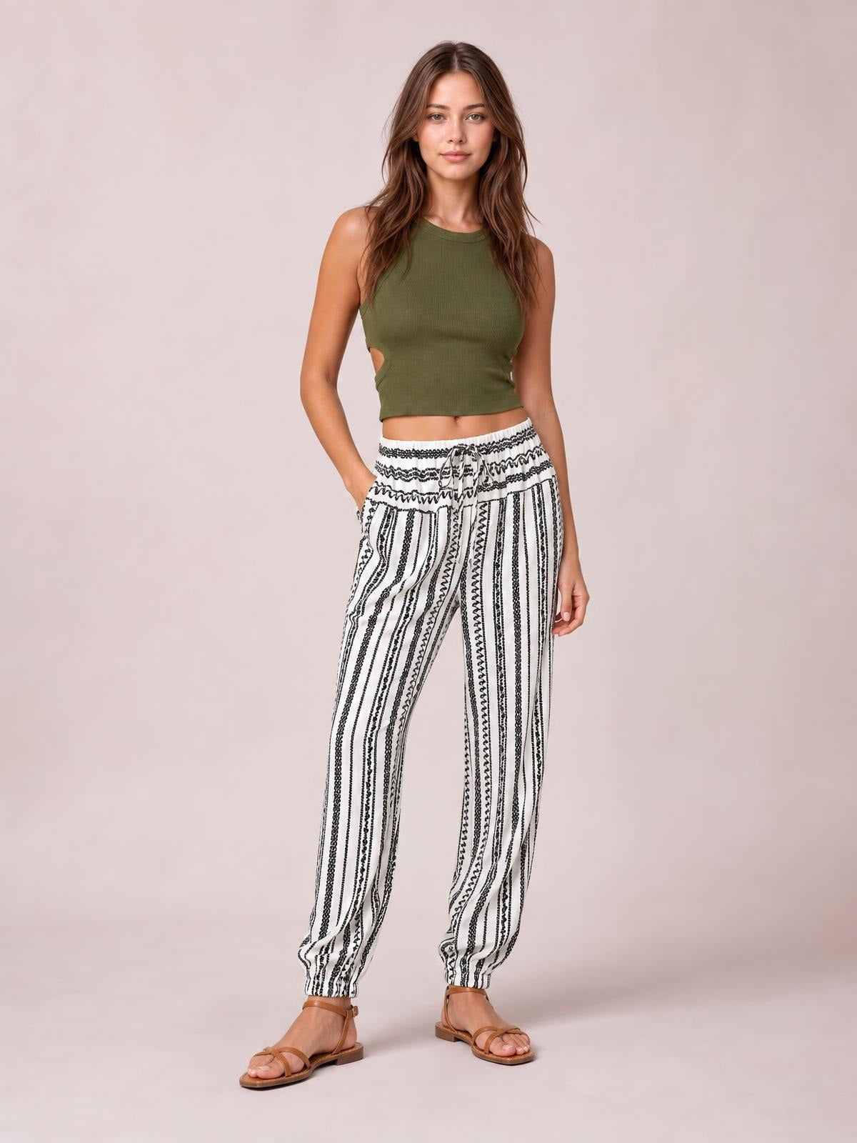Model poses with one hand in the pocket, wearing white printed elastic-waist pants with black vertical stripes, a drawstring waist, and tapered ankles.