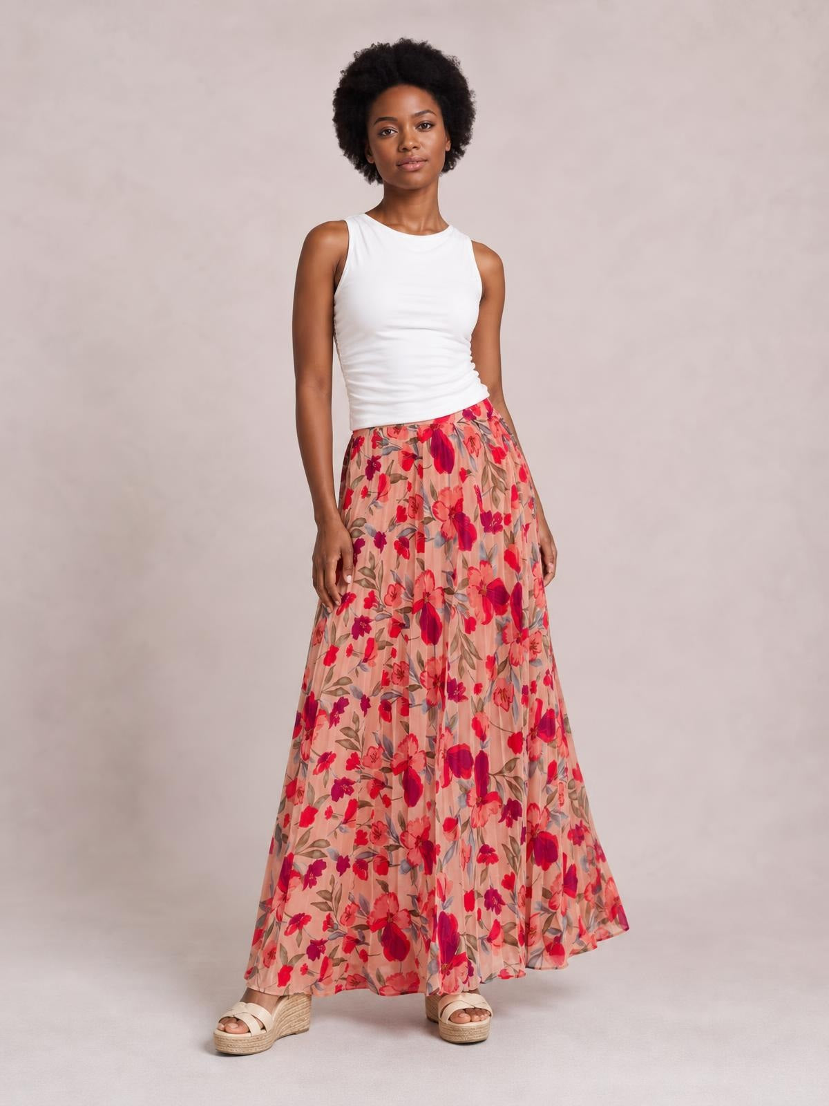 Model stands with one leg slightly forward in a flowing peach floral maxi skirt featuring red blooms, vertical pleats, an elastic waist, and a softly flared silhouette.