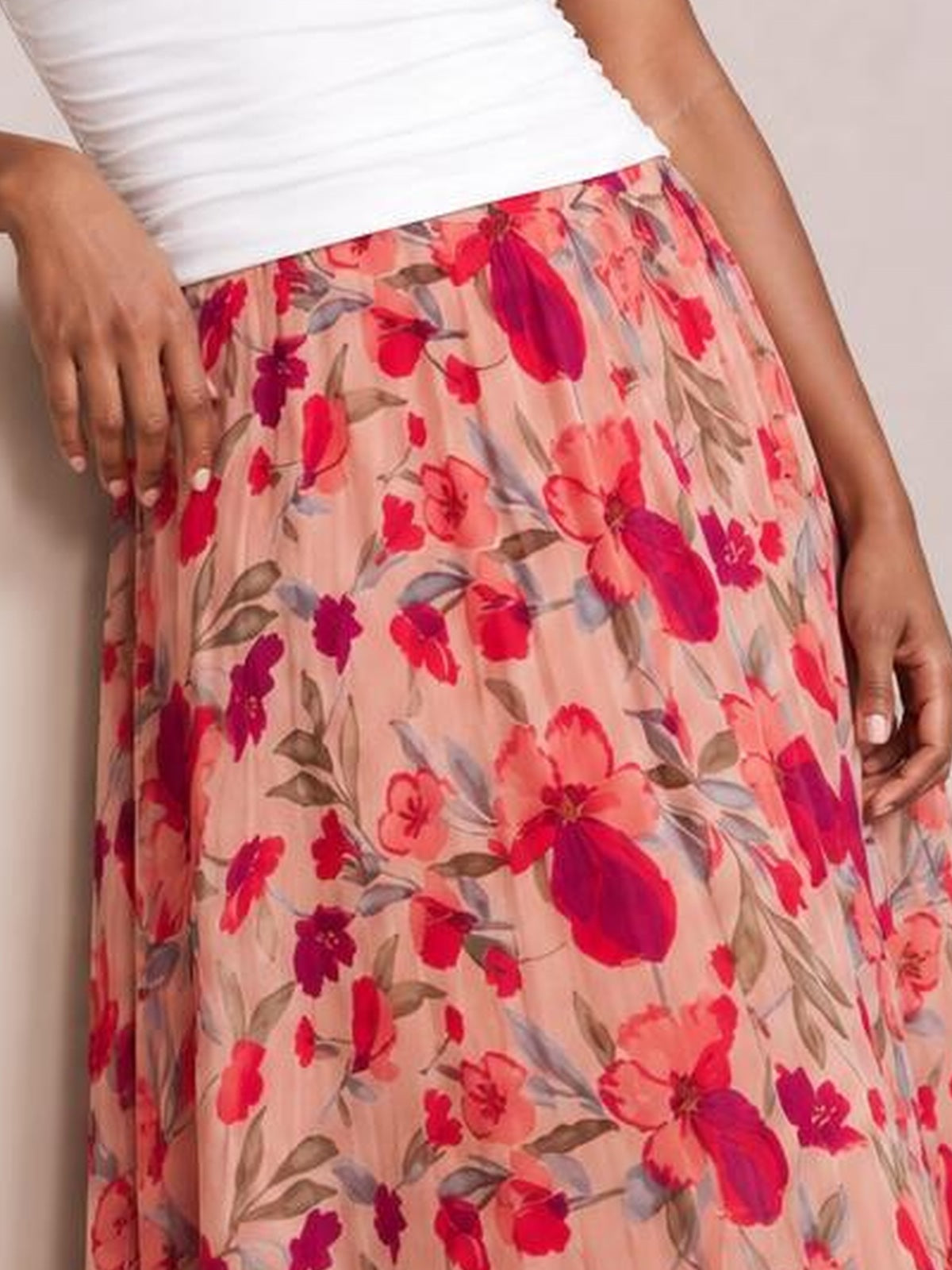 A model wears a flowy pink floral maxi skirt with an elastic waist, soft vertical pleats, and a relaxed silhouette, with hands resting at her sides.