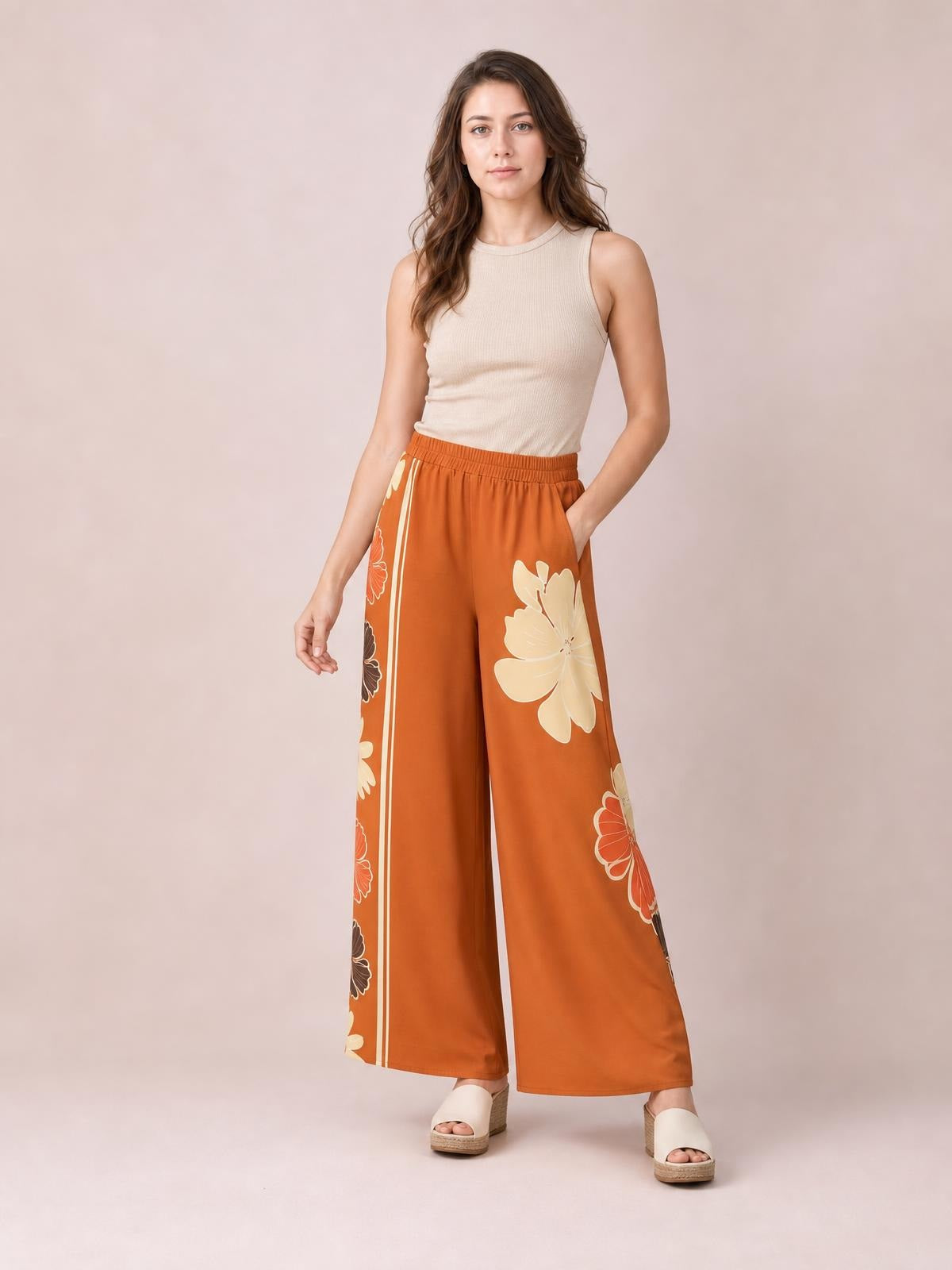 Model poses in printed rust-orange elastic waist wide leg pants with oversized cream floral motifs and straight side stripes.