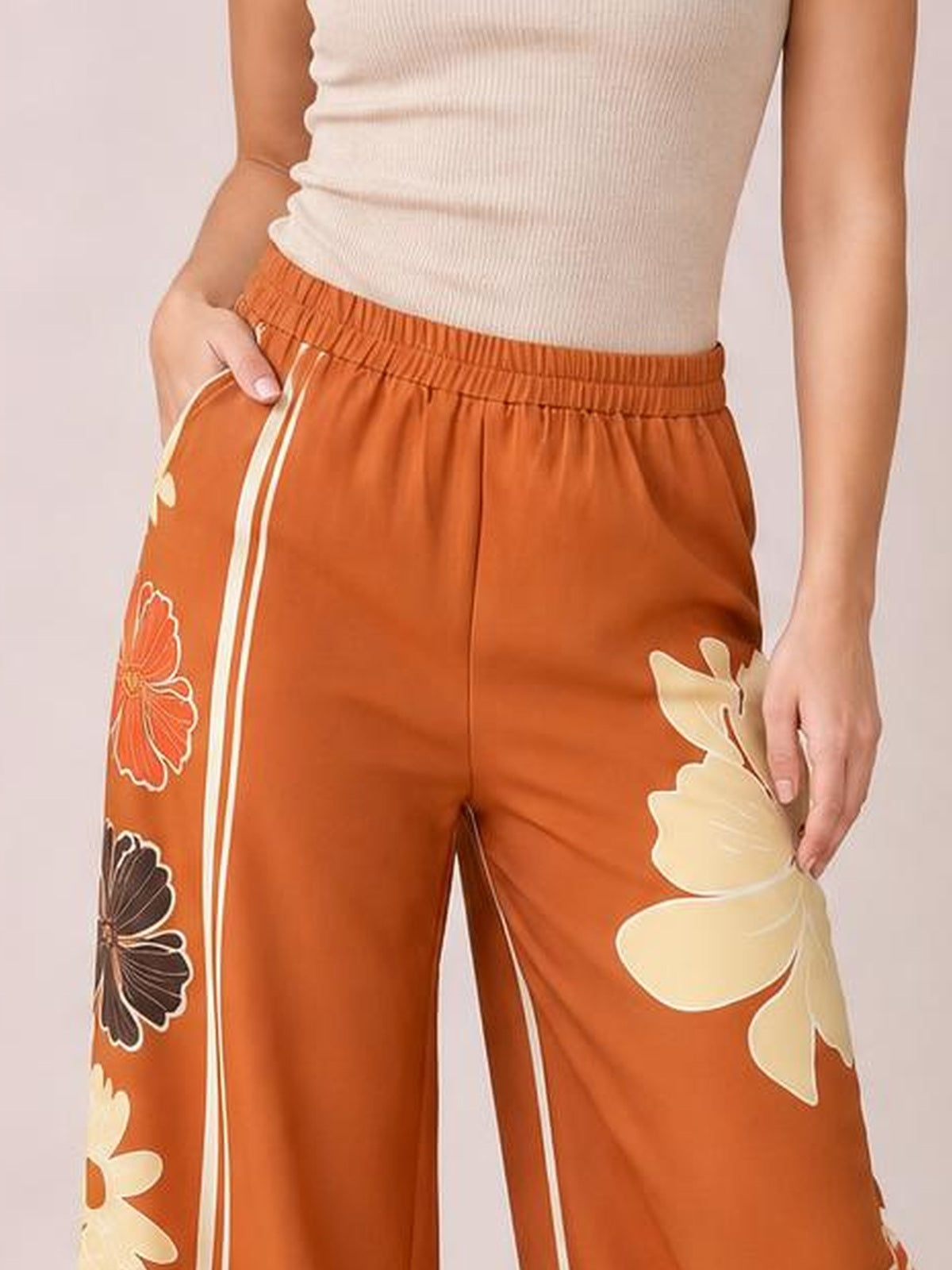 Model wears rust-orange printed elastic waist wide leg pants with cream and black floral motifs, white side stripes, and a relaxed silhouette.
