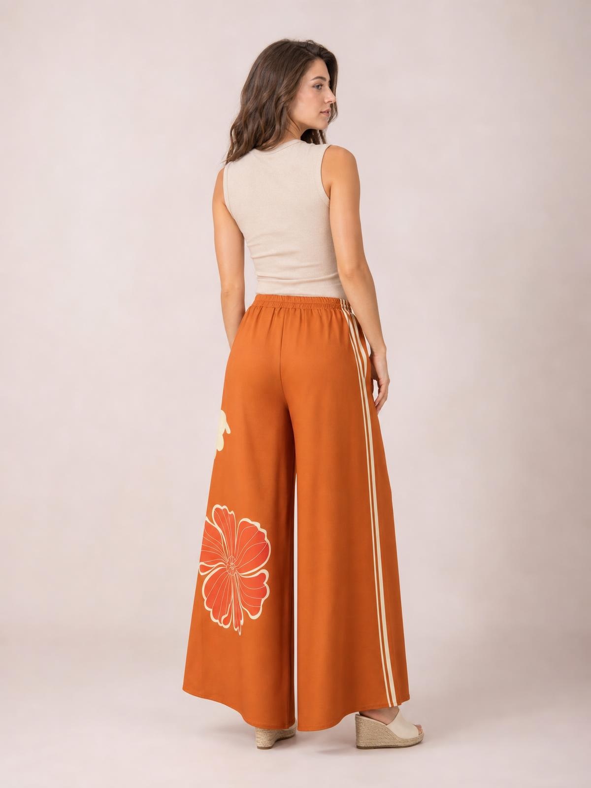 Model shown from behind wears rust-orange printed elastic waist wide leg pants with white floral motifs and side stripes in a flowing, full-length silhouette.