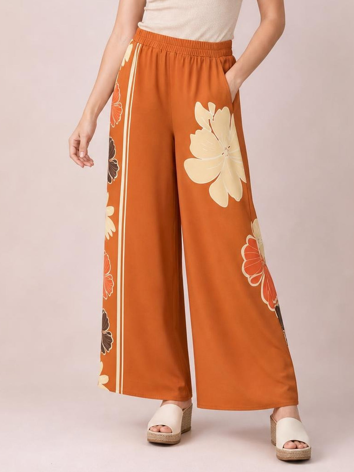 Model wearing rust-orange printed elastic waist wide leg pants with cream floral accents and side stripes, posing with one hand in a pocket.