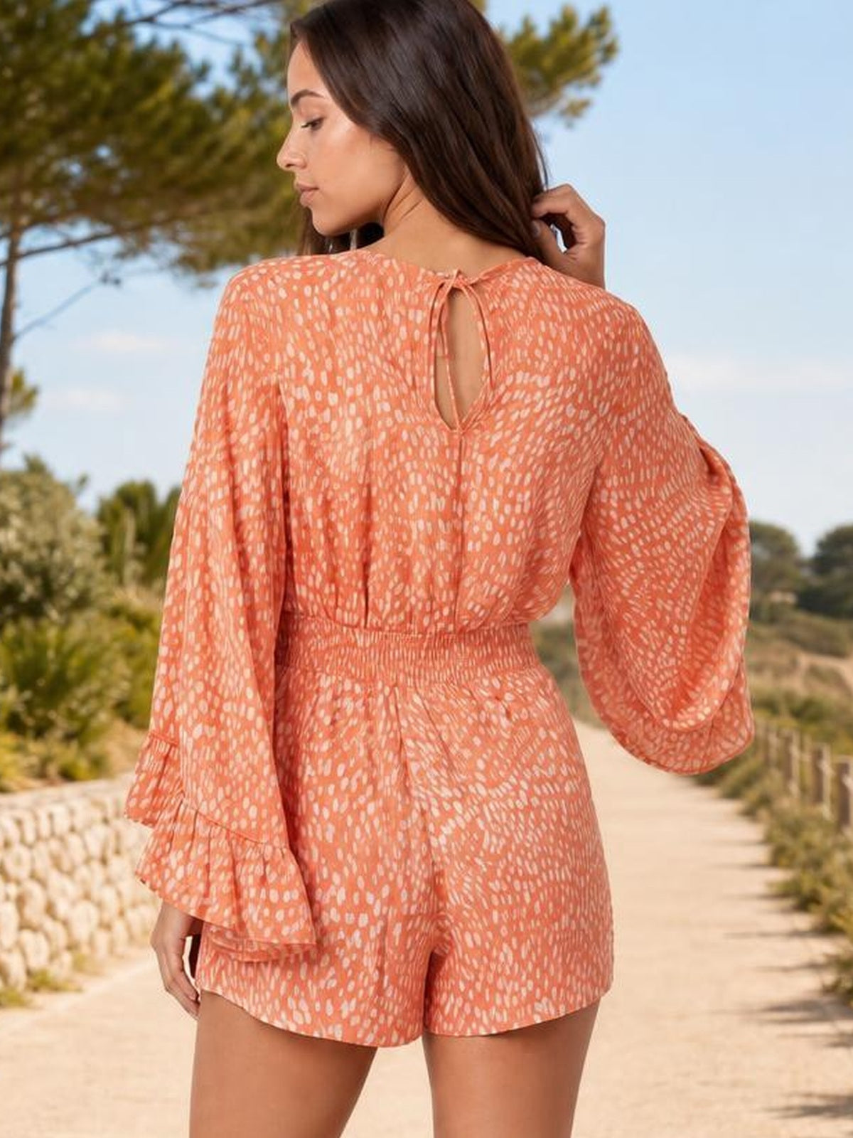 A model wears a coral printed romper with long flare sleeves, a smocked waist, relaxed shorts silhouette, and a tied keyhole back while touching her hair.