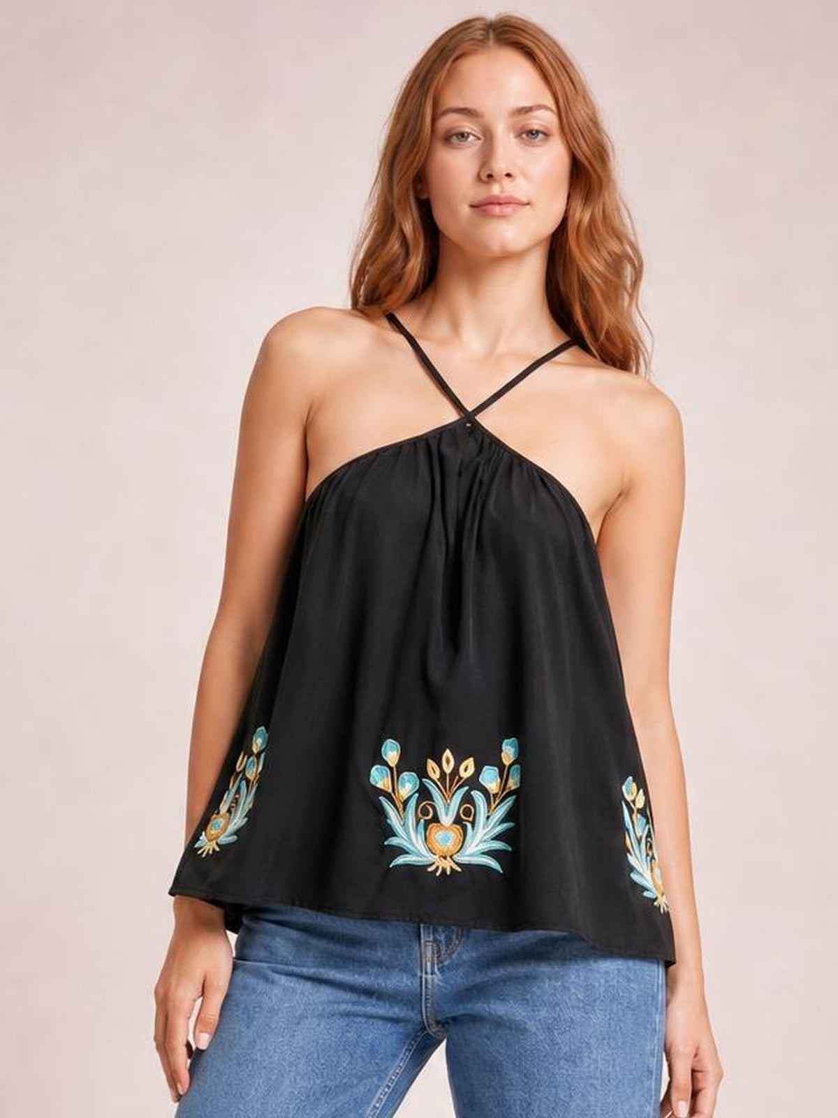 Model poses in a black printed Grecian neck cami with crossed straps, gathered neckline, and a loose flowy silhouette with floral embroidery.