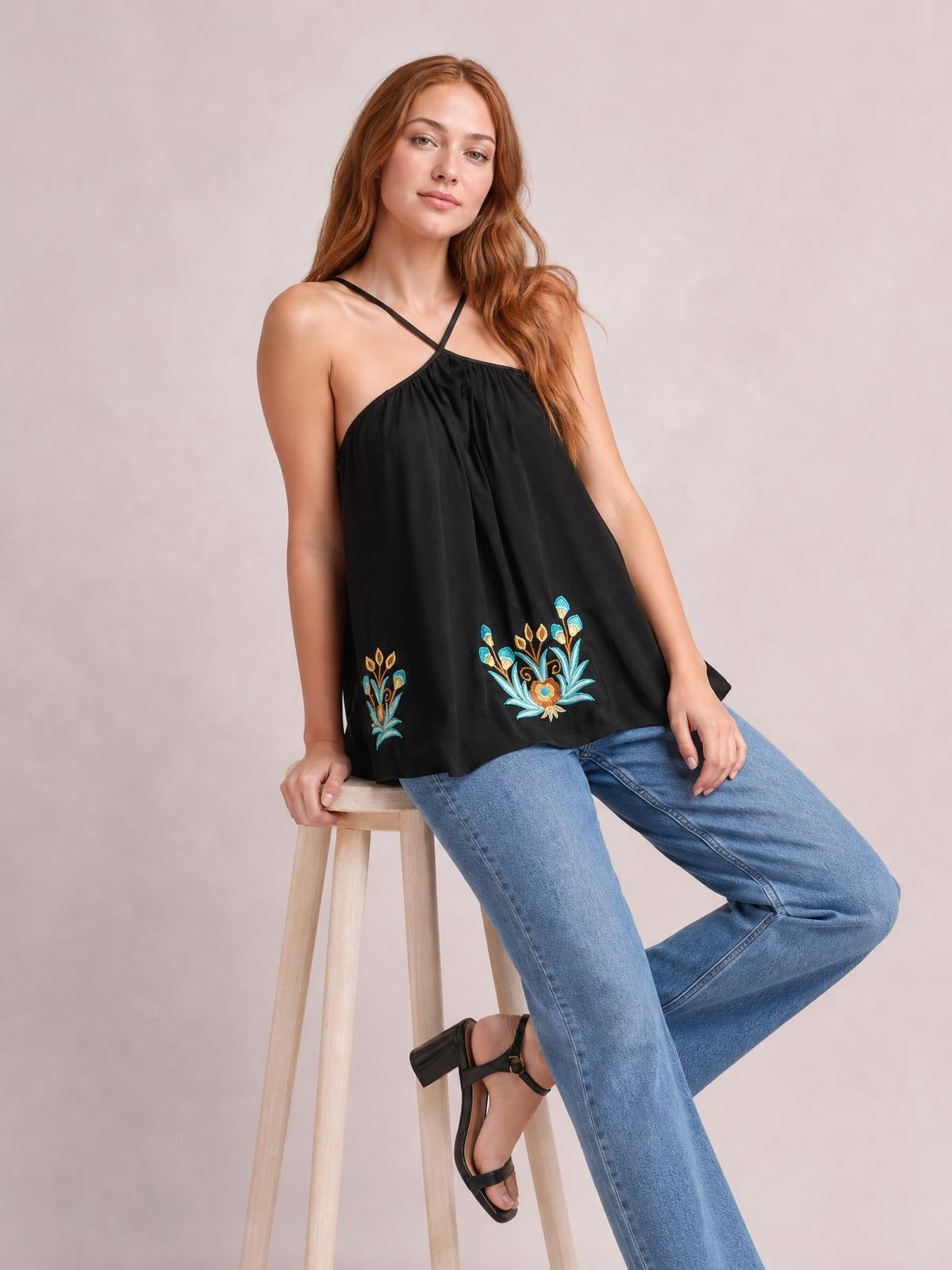 A model sits wearing a black printed Grecian neck cami with crisscross straps, a gathered neckline, and a loose flowing silhouette with floral motifs near the hem.