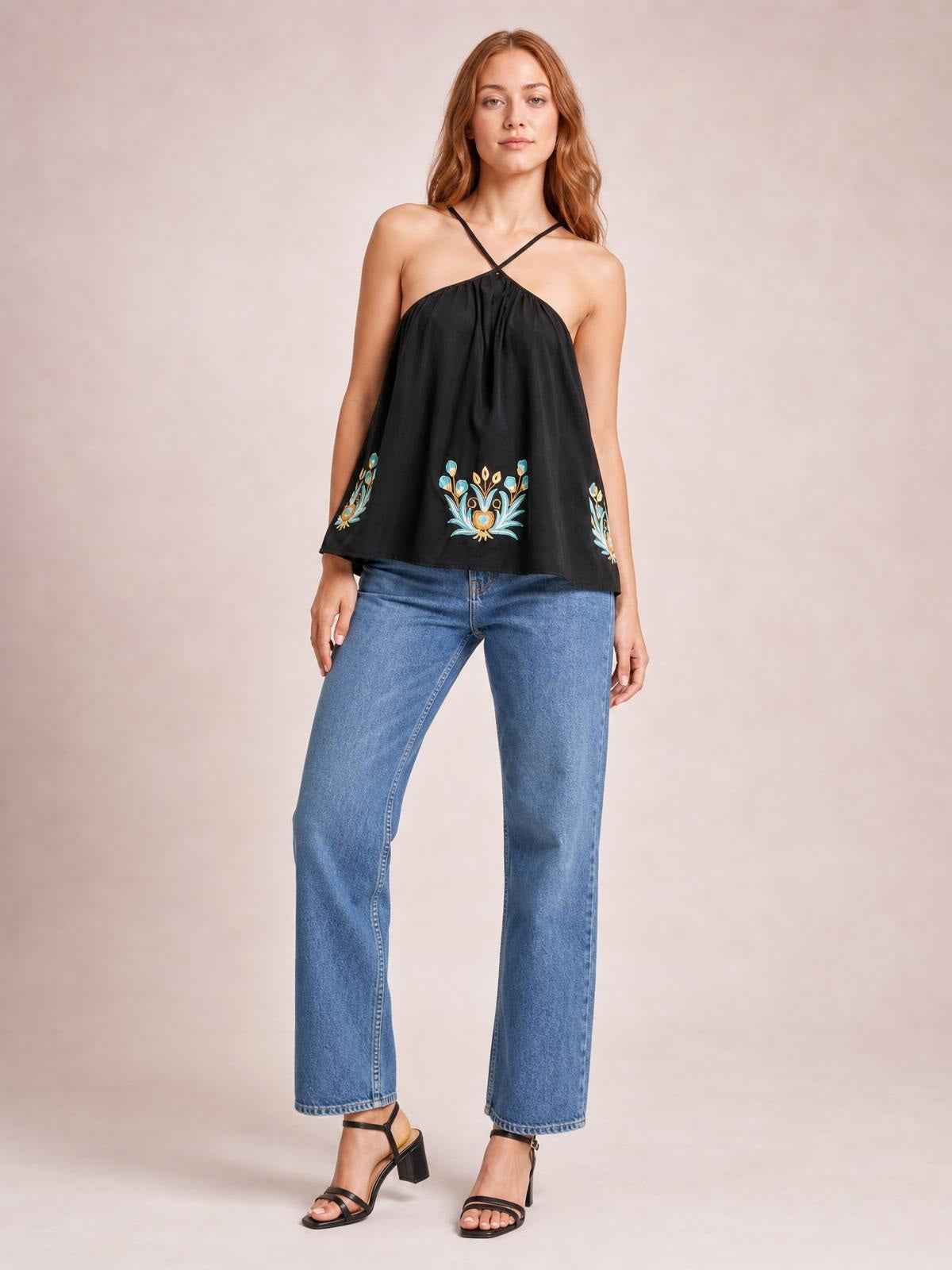 A model stands in a black printed Grecian neck cami with crossed straps, a loose flowy silhouette, and colorful floral motifs along the hem.
