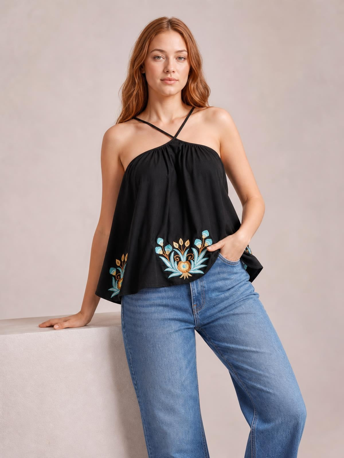 Model poses in a black printed Grecian neck cami with crossed straps, gathered neckline, and a loose flared silhouette with colorful floral motifs.