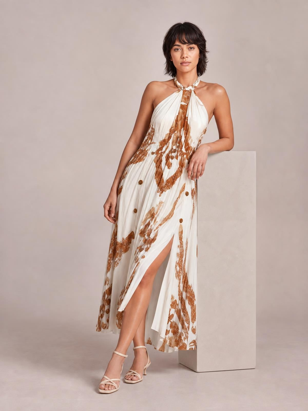 Model in a white and brown printed halter neck maxi dress with a flowing silhouette, gathered neckline, and high side slit, leaning against a pedestal.