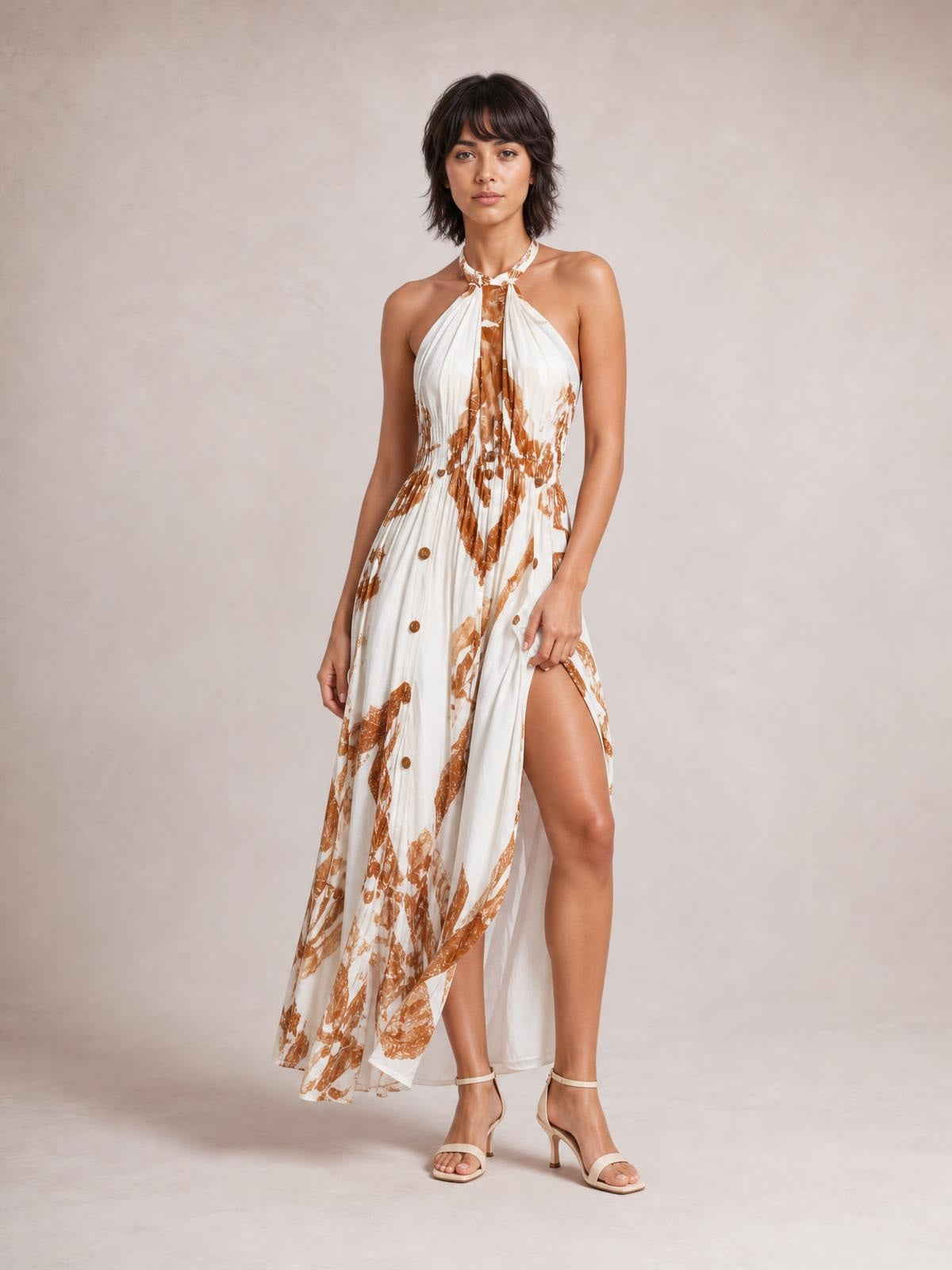 A model poses in a white and brown printed halter neck maxi dress with a gathered waist, button-front detail, flowing silhouette, and high side slit.