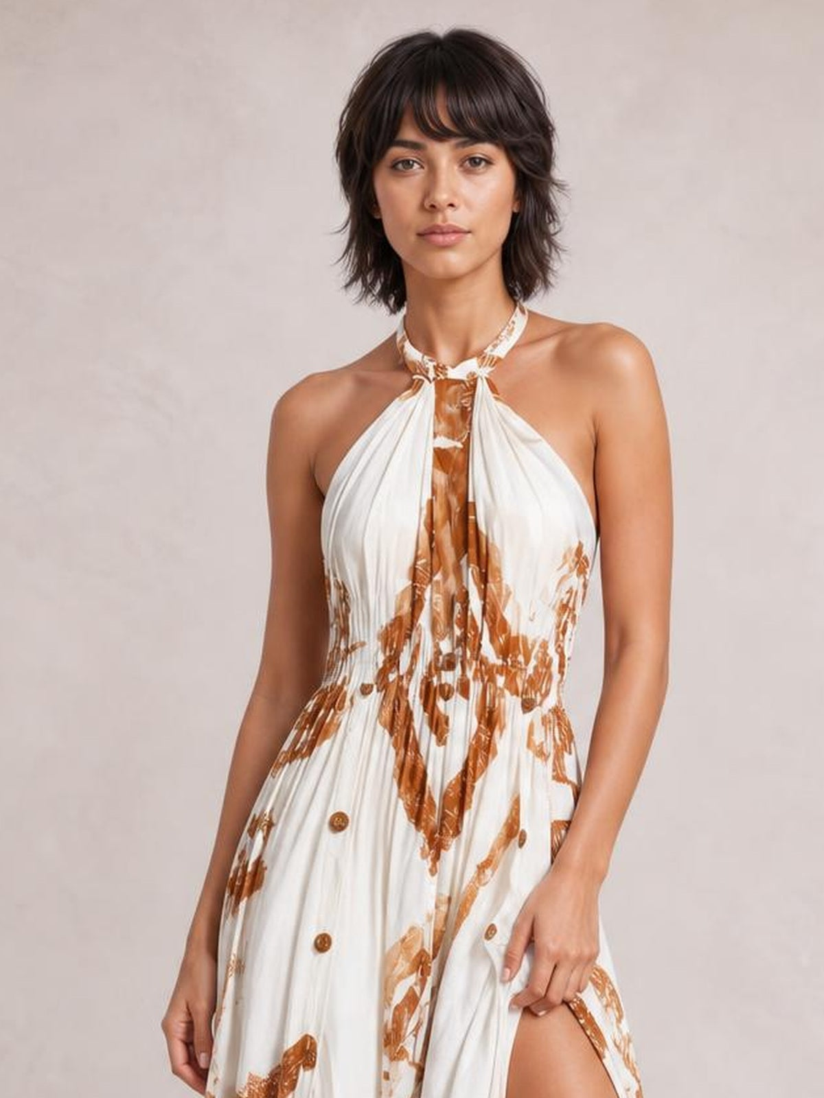 A model poses in a white and brown printed halter neck maxi dress with a gathered bodice, flowing silhouette, and thigh-high slit.