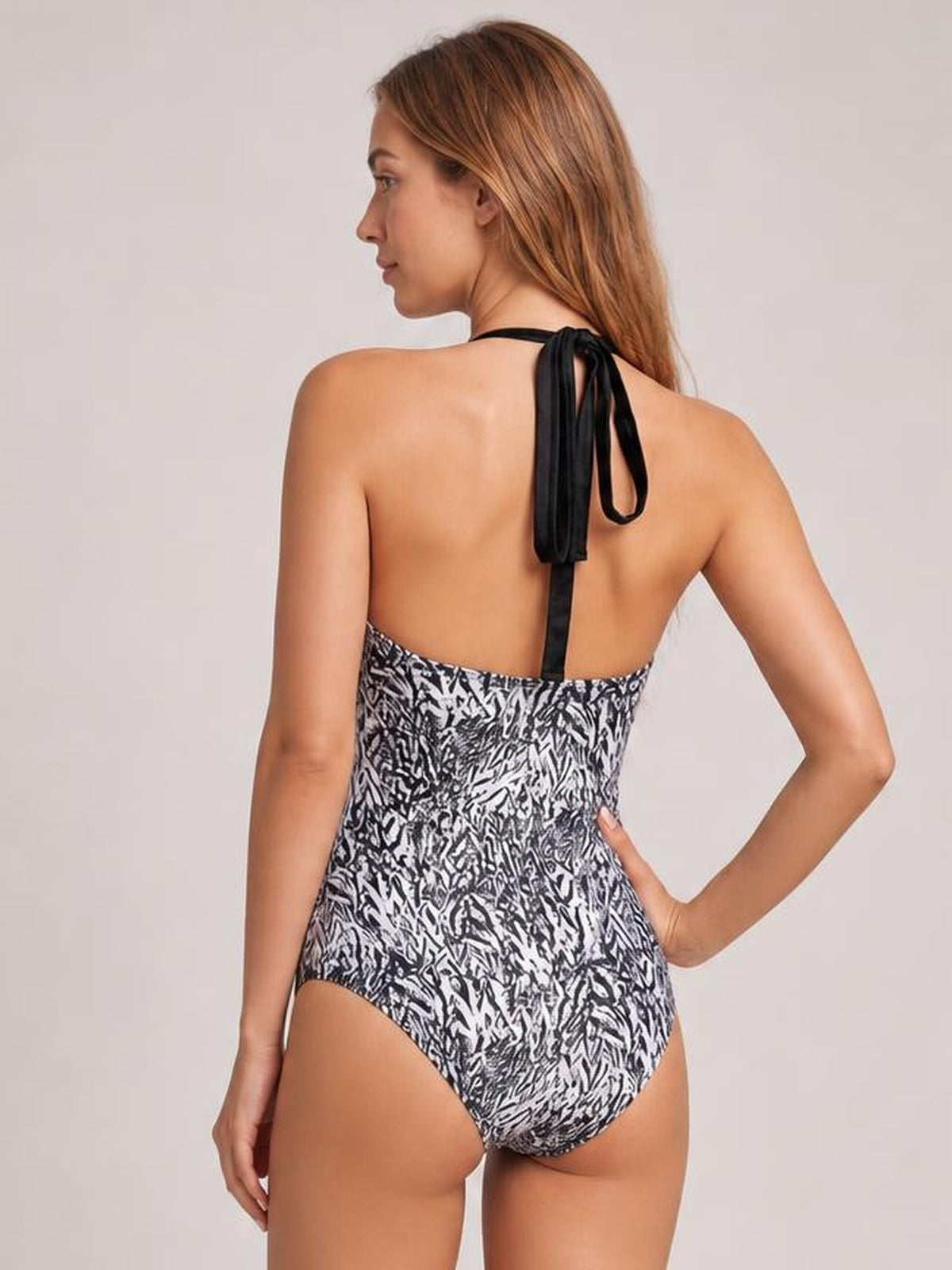Model poses with one hand on her hip, showing the back of a black-and-white printed halter-neck one-piece swimsuit with tied straps and a high-cut silhouette.