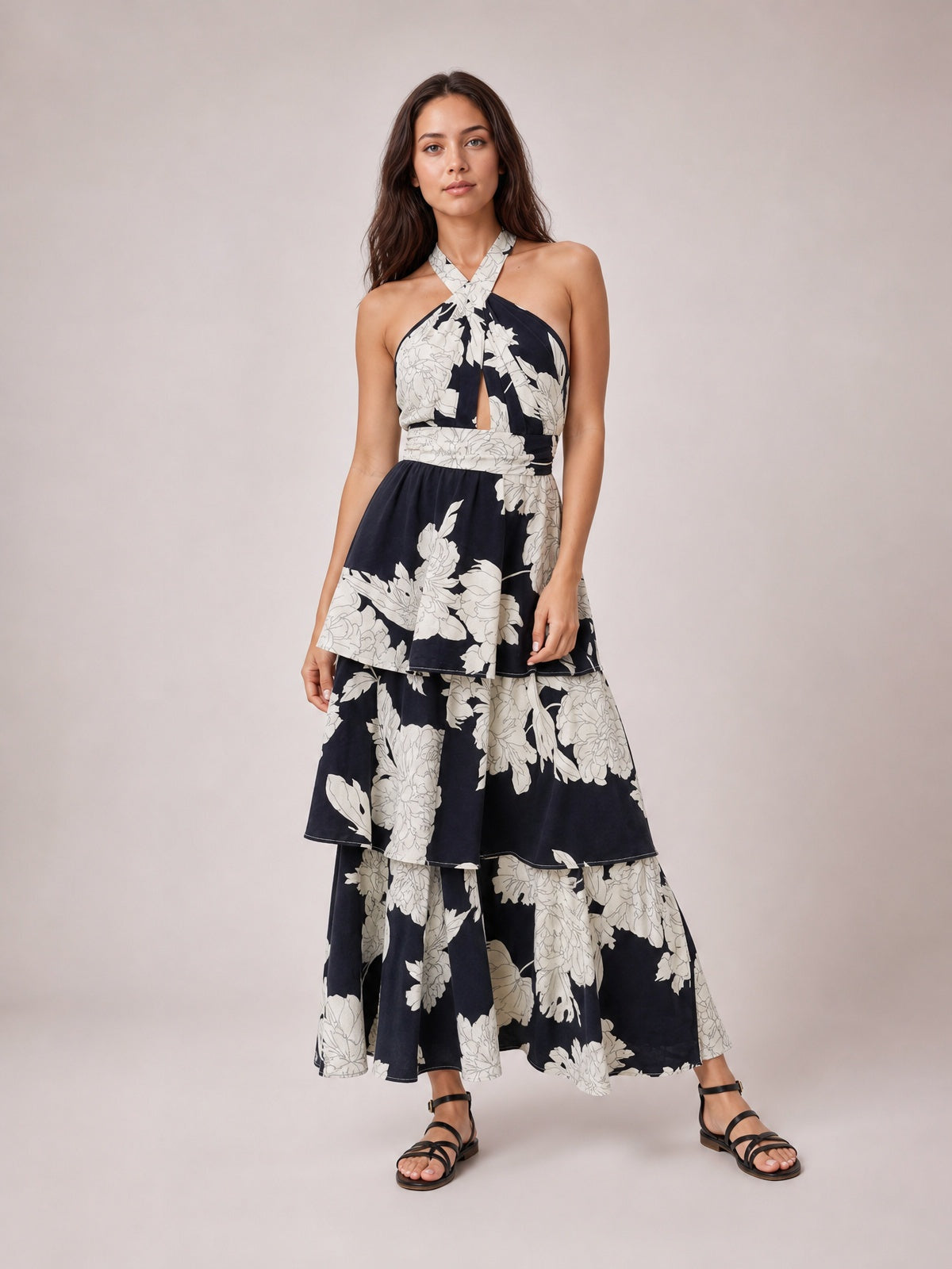 Model stands with one leg angled in a black floral halter maxi dress featuring a crisscross neckline, center cutout, defined waist, and tiered skirt.