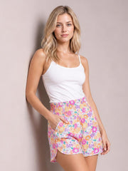 A woman wearing a white camisole top and colorful floral shorts, posing against a solid light beige wall.