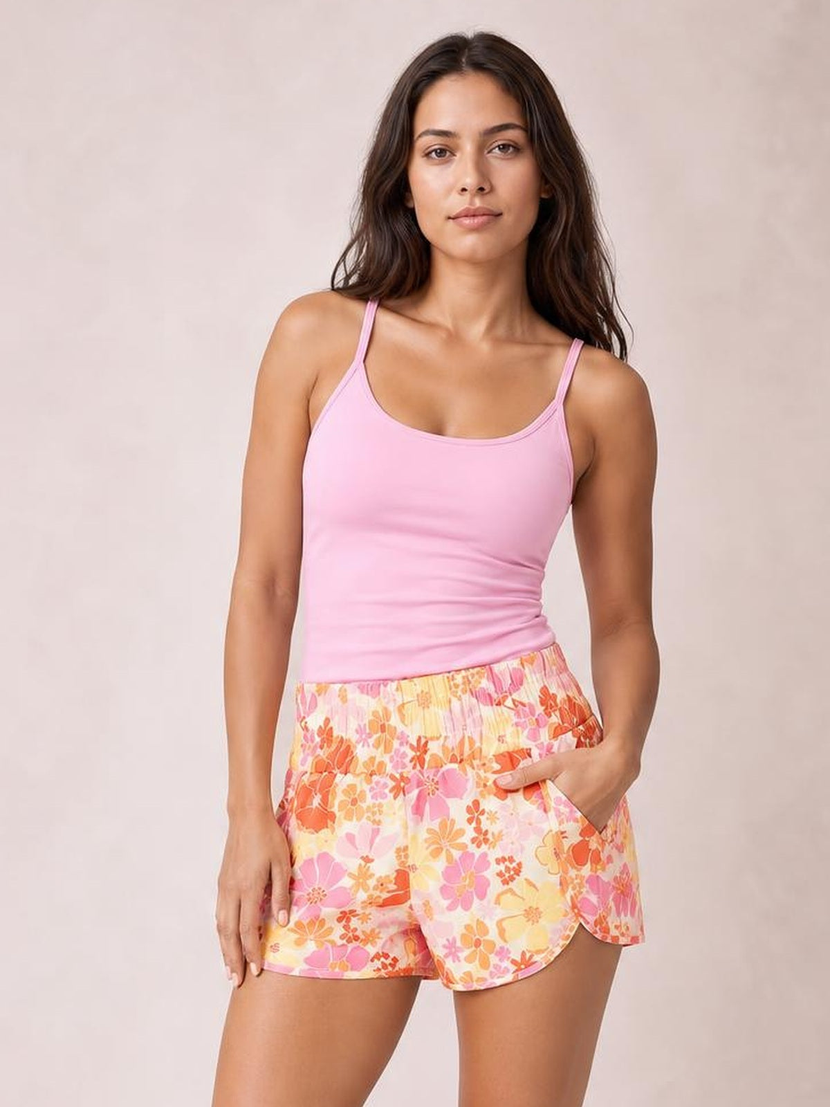 A woman modeling a light pink ribbed tank top and high-waisted shorts with a vibrant orange, pink, and yellow floral print, set against a neutral beige studio background.