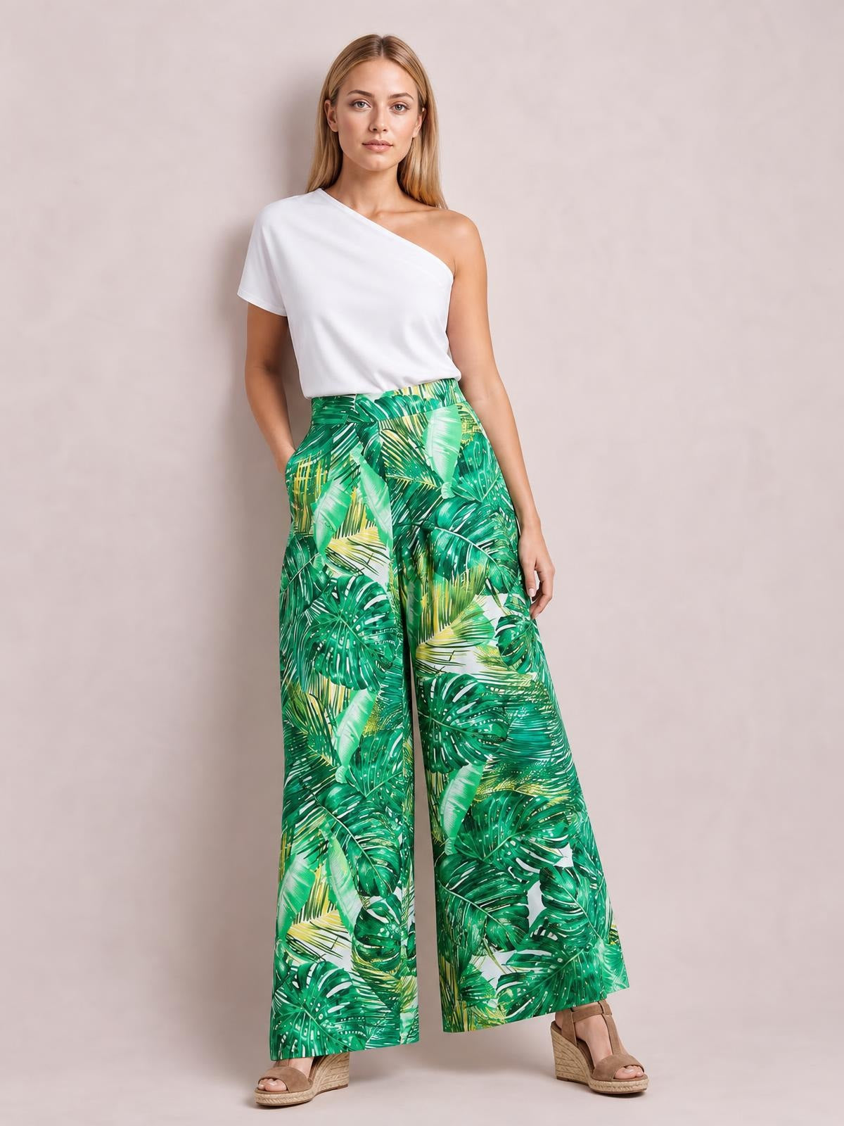 Model poses in green tropical-leaf printed high waist wide leg pants with a flowing silhouette, wide waistband, and side pockets.