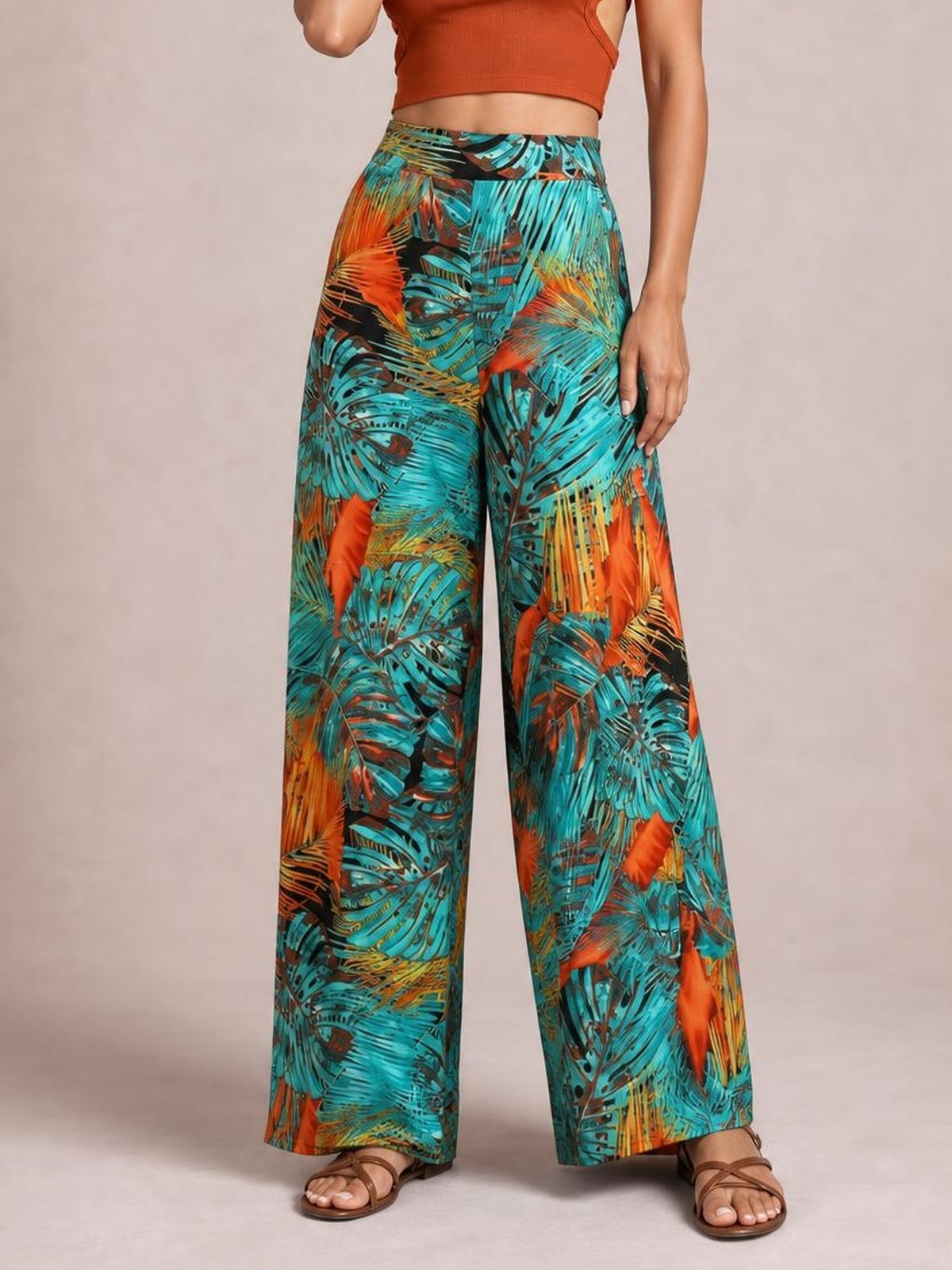 A model stands with one leg slightly forward in colorful printed high-waist wide-leg pants featuring turquoise, orange, and black tropical leaf motifs.