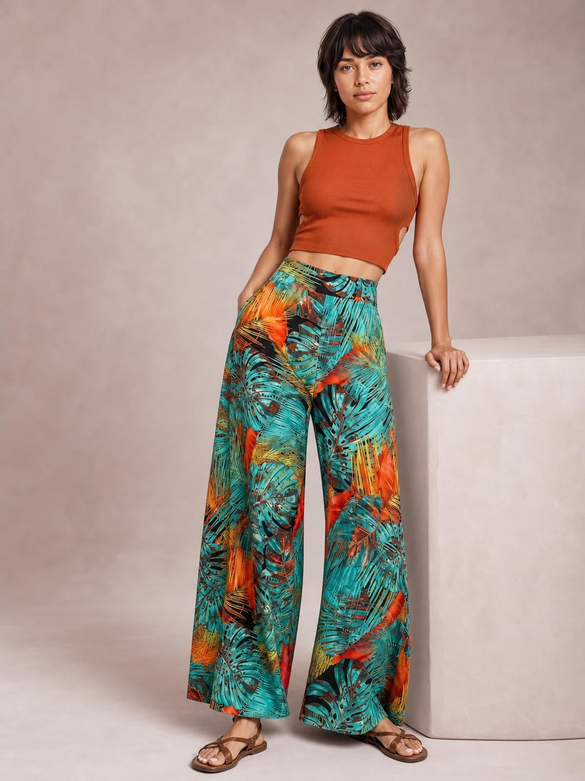 Model poses in printed high waist wide leg pants with a vibrant tropical leaf pattern in turquoise, orange, and black, featuring a flowing silhouette.