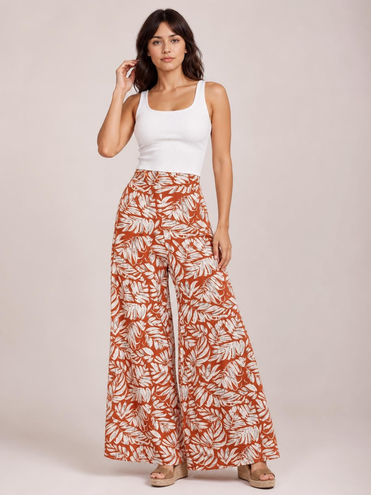 Model poses in rust-brown printed high waist wide leg pants with a white leaf pattern, flowing full-length silhouette, and a fitted waistband.