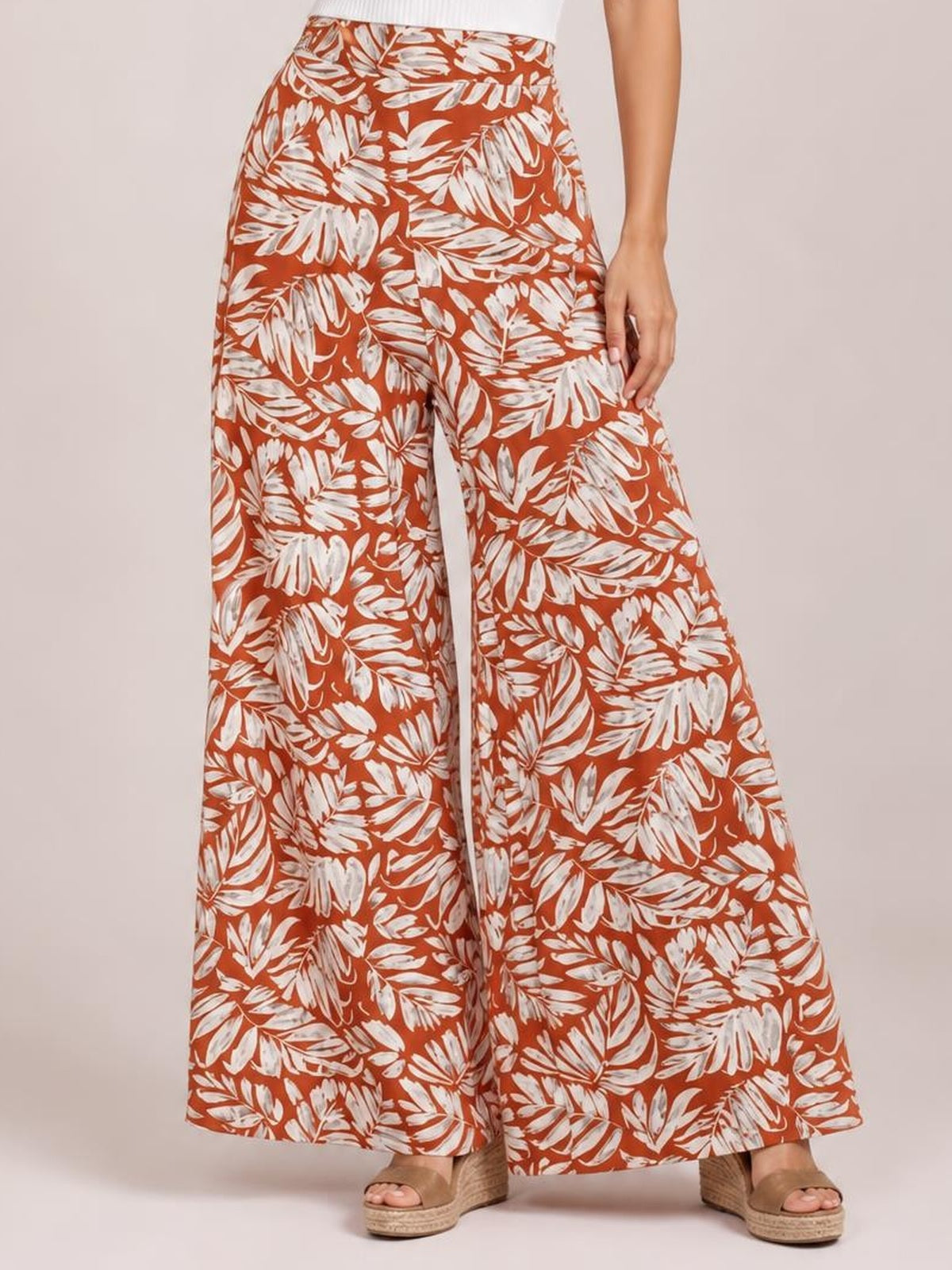 Model wearing rust-orange printed high waist wide leg pants with an opaque white botanical pattern, flowing full-length silhouette, and wide hems.