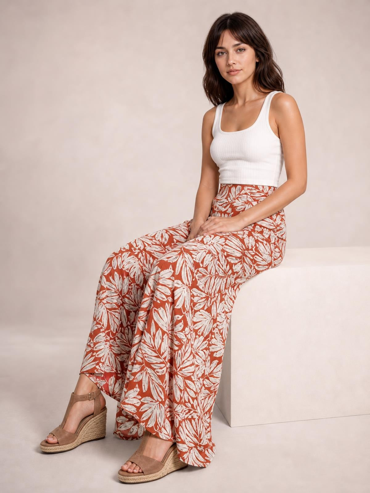 Model seated in printed rust and white high waist wide leg pants with a flowing silhouette and bold botanical pattern, paired with a white tank top.