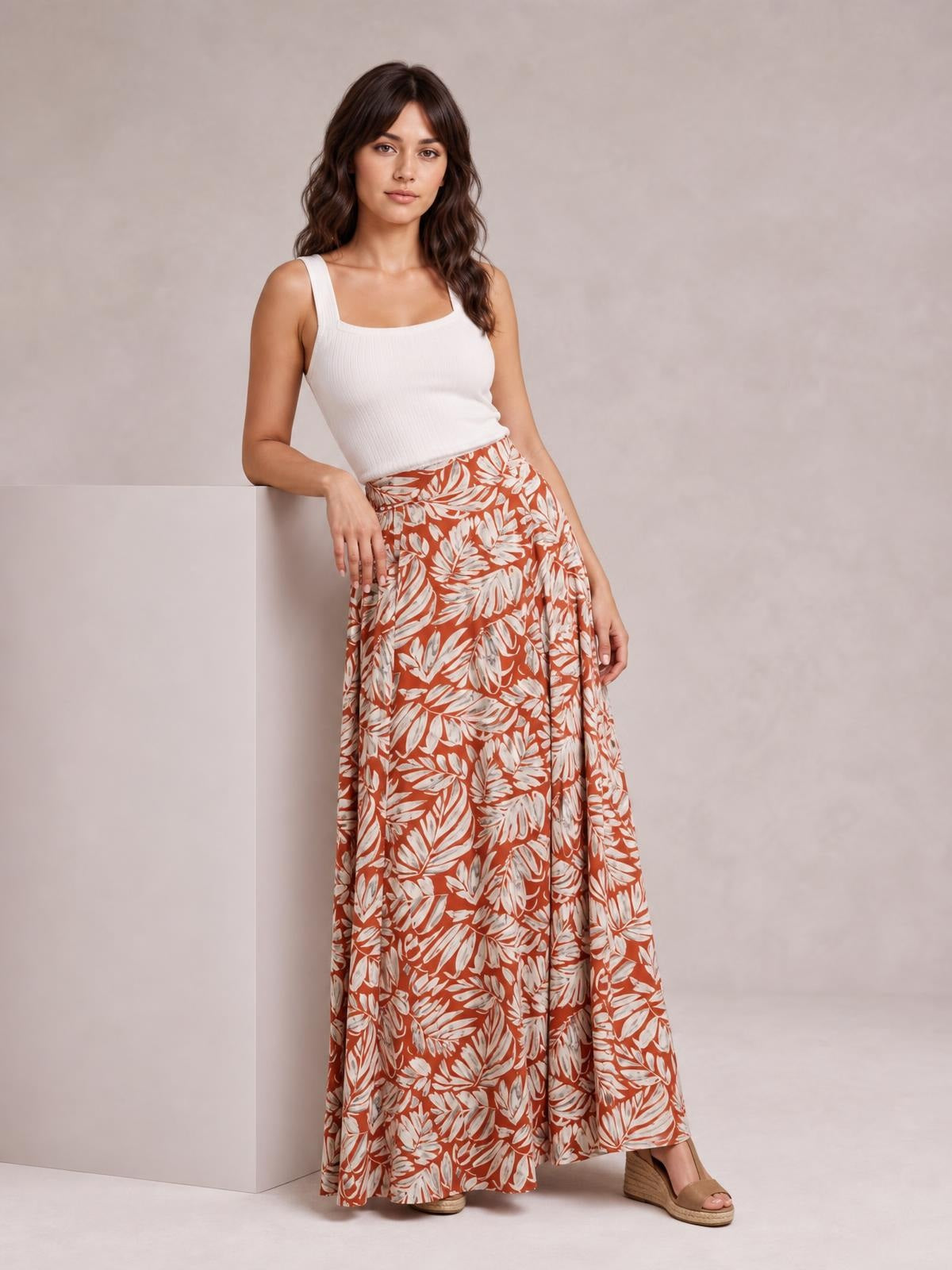 A model leans against a block wearing rust-orange printed high-waist wide-leg pants with a flowing white botanical pattern and relaxed silhouette.