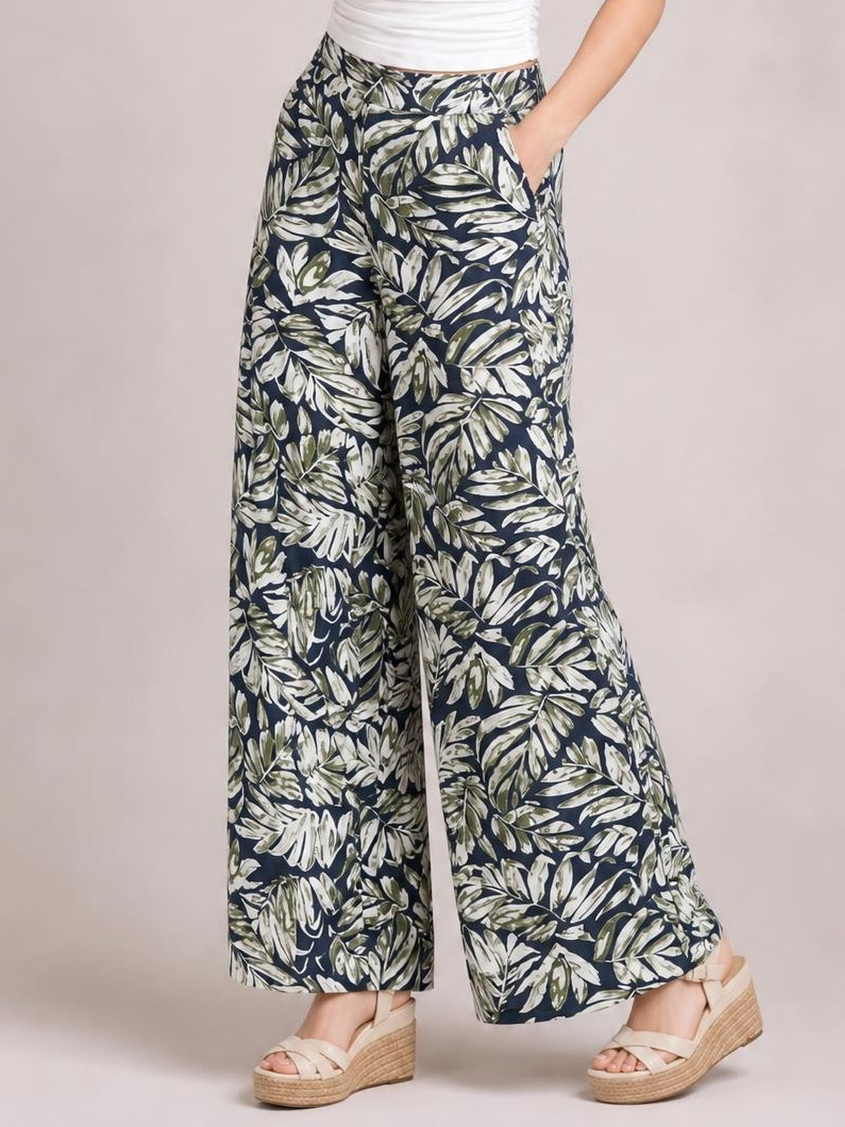 Model poses in printed high waist wide leg pants with a navy base, leafy cream and olive pattern, side pockets, and a flowing full-length silhouette.