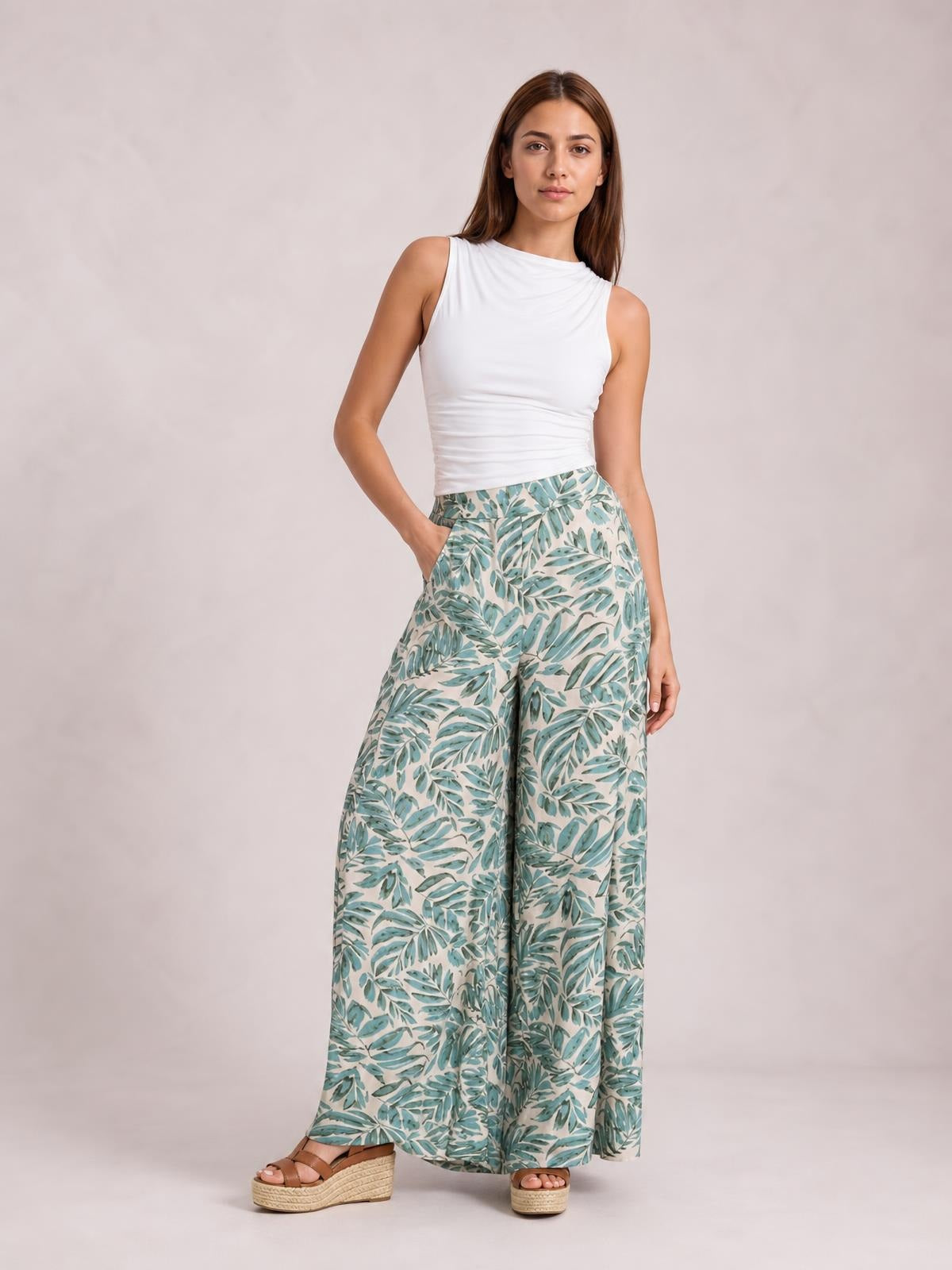 Model poses in printed high waist wide leg pants with a flowing teal and cream leaf pattern, wide silhouette, and side pockets, paired with a white sleeveless top.