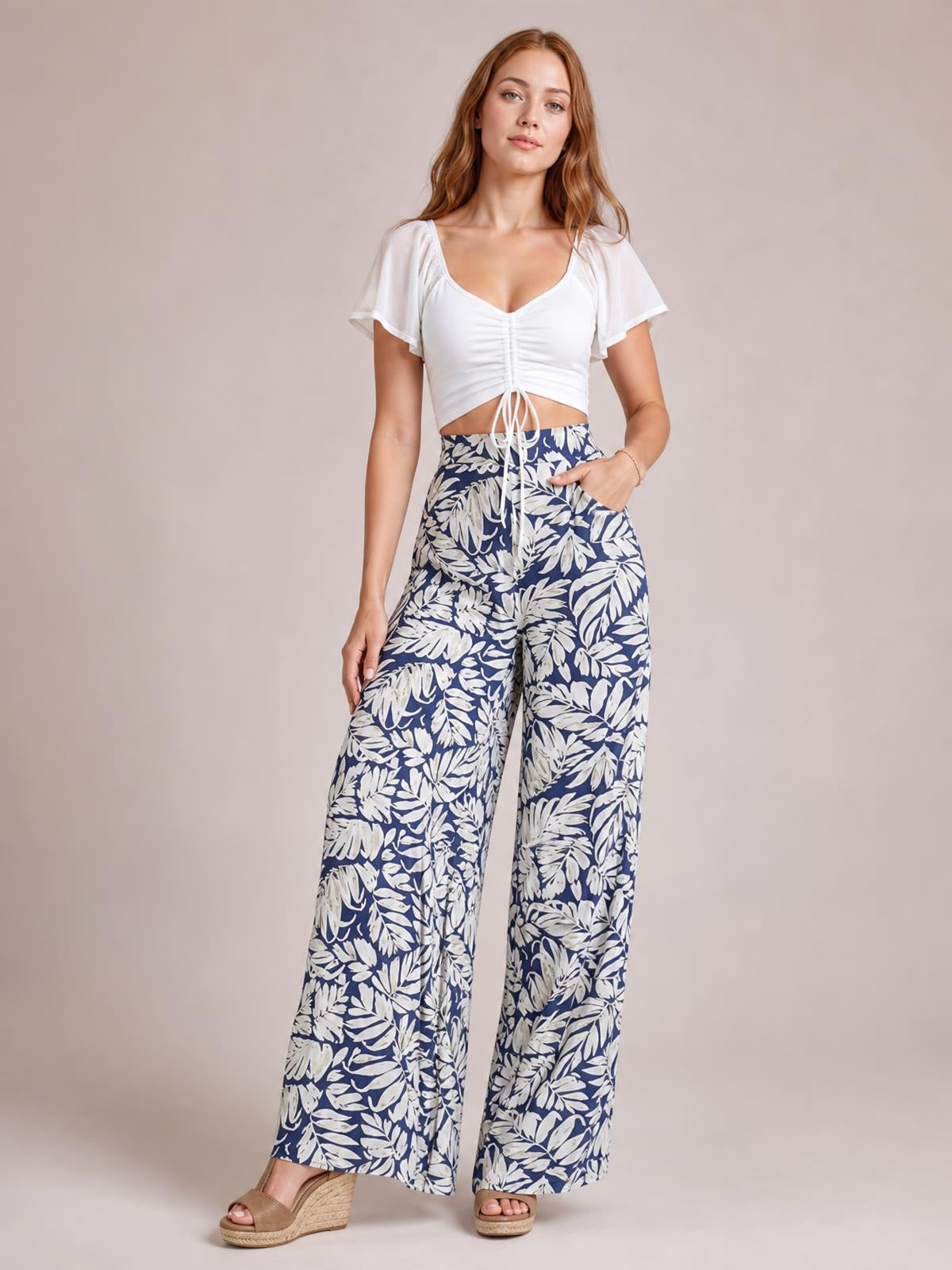 A model stands with one hand in a pocket, wearing opaque navy and white leaf-print high-waist wide-leg pants with a flowing silhouette.