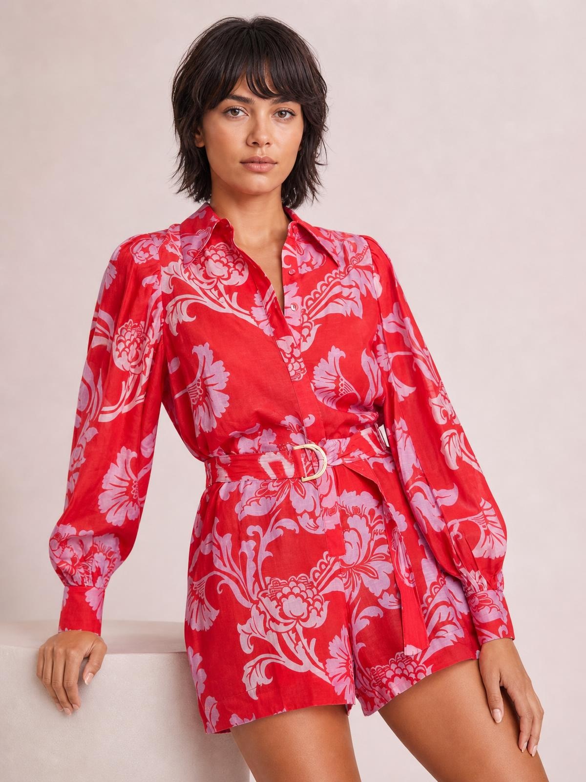 Model poses in a red and lavender printed long sleeve romper with a belted waist, button-front neckline, relaxed silhouette, and cuffed sleeves.