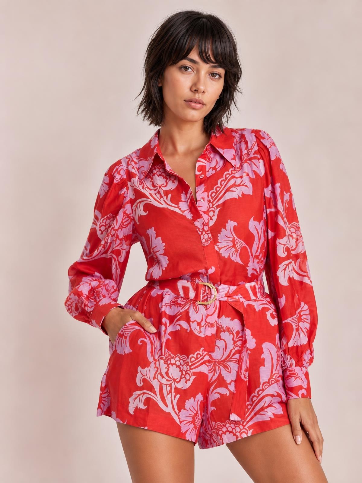 Model poses with one hand in a pocket wearing a red floral-print long-sleeve belted romper with a button-front shirt collar and relaxed shorts silhouette.
