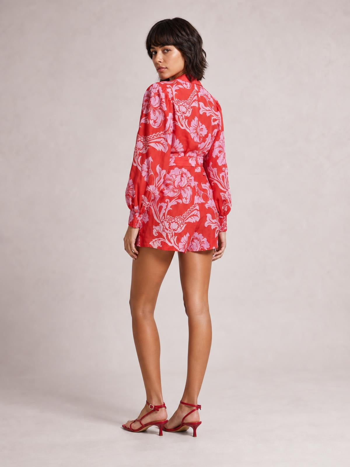 Model poses turned away in a red and pink printed long-sleeve romper with a belted waist, buttoned front, balloon sleeves, and relaxed shorts silhouette.