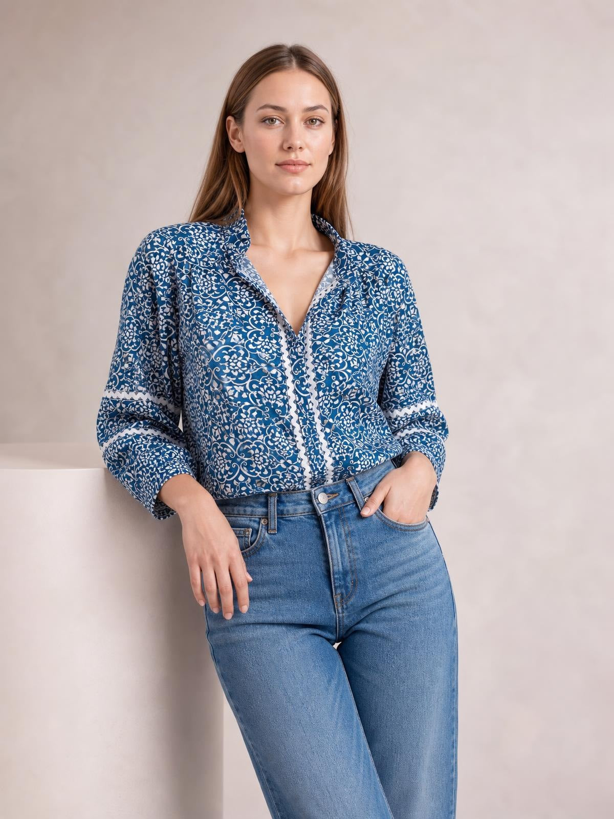 A model wears a blue-and-white printed mock neck long sleeve blouse with a button-front construction, relaxed silhouette, and cuffs, posing with hands in jeans pockets.