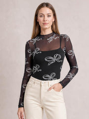 Model wears a fitted black sheer mesh T-shirt with a mock neck, long sleeves, and white bow print, posing with one hand in her jeans pocket.
