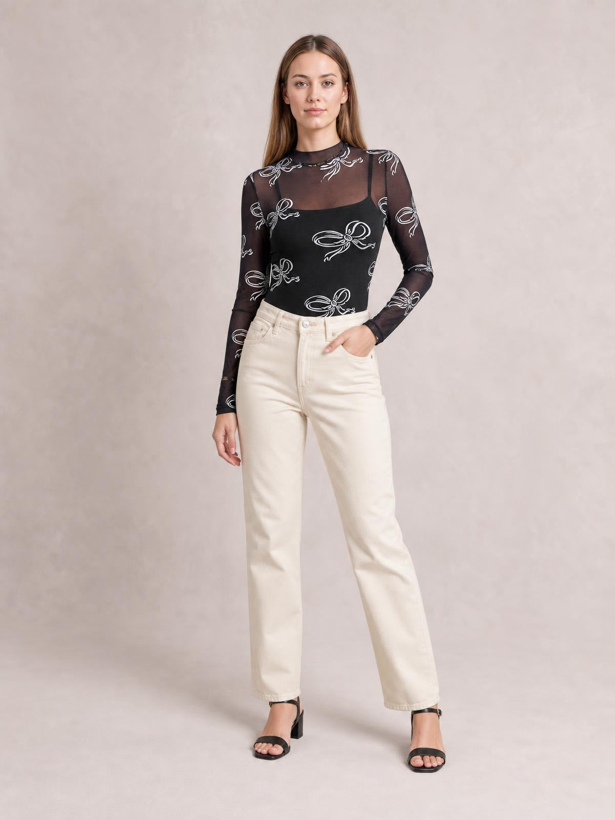 Model poses in a black sheer mesh mock neck long sleeve T-shirt with white bow print, fitted silhouette, and a hand in her pocket.