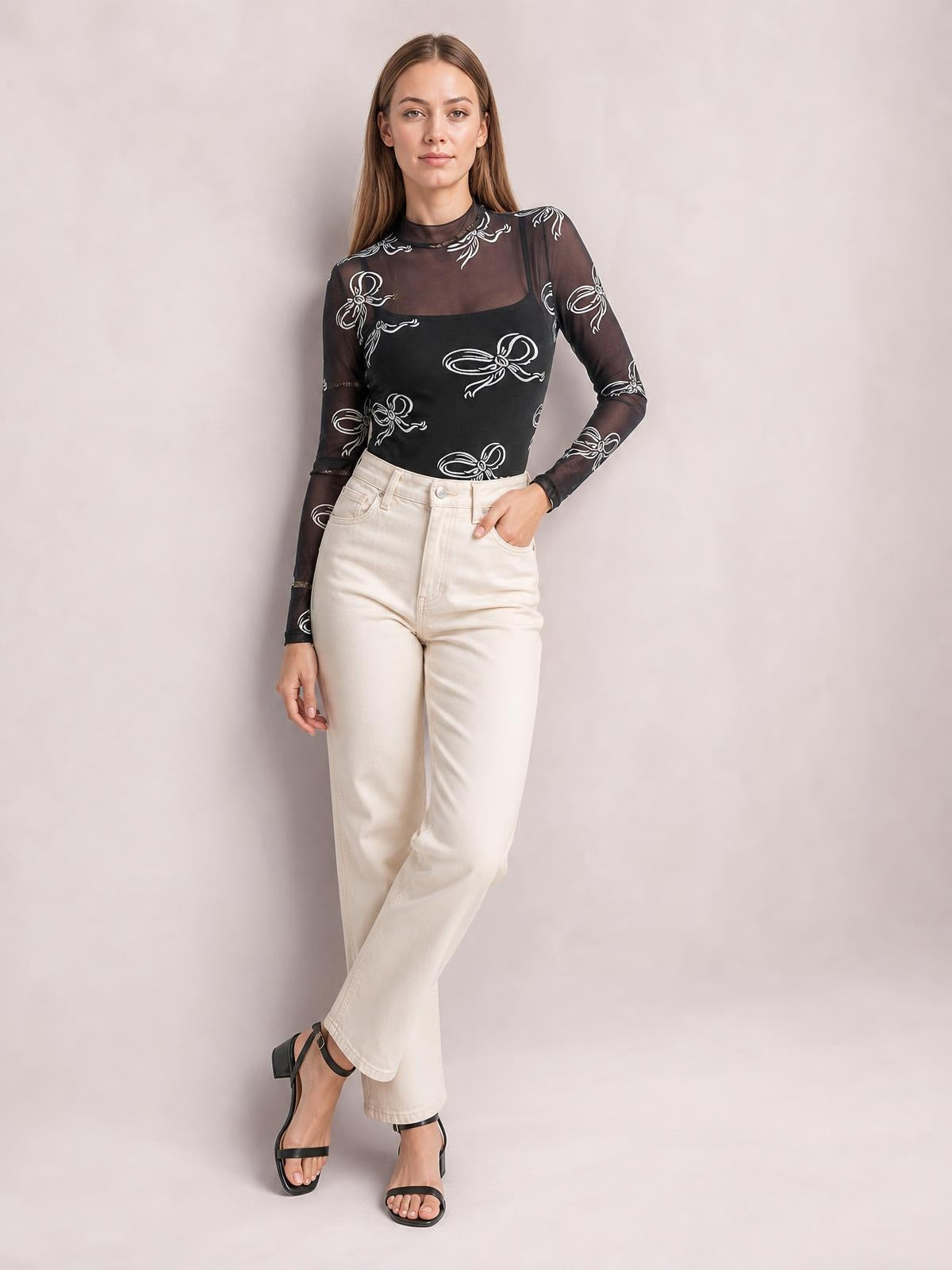 Model wears a black printed mock neck long sleeve mesh T-shirt with sheer construction, a fitted silhouette, and white bow motifs.
