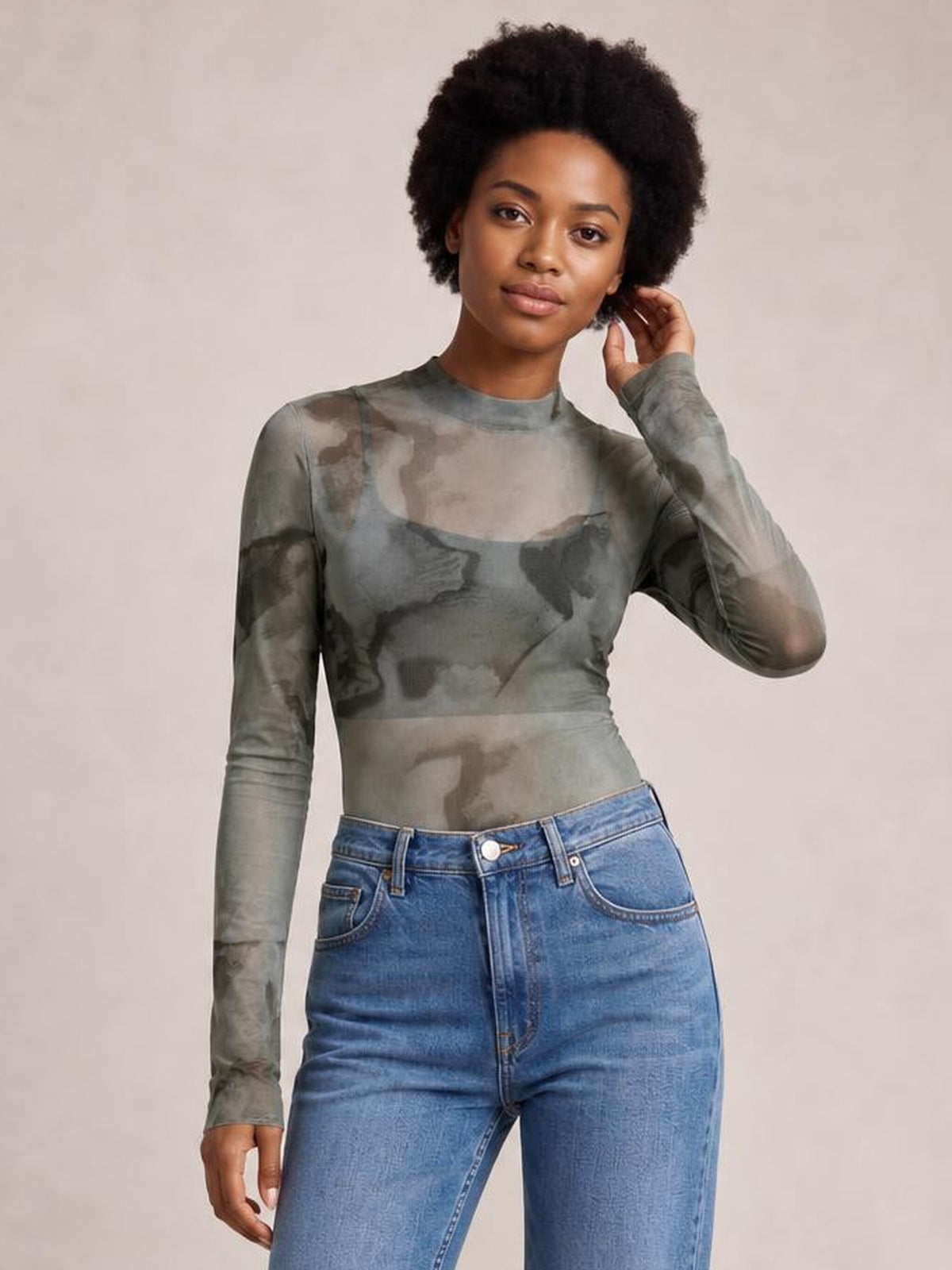 Model poses in a fitted gray-green printed mock neck long sleeve mesh T-shirt with sheer construction and an abstract pattern, touching her hair.