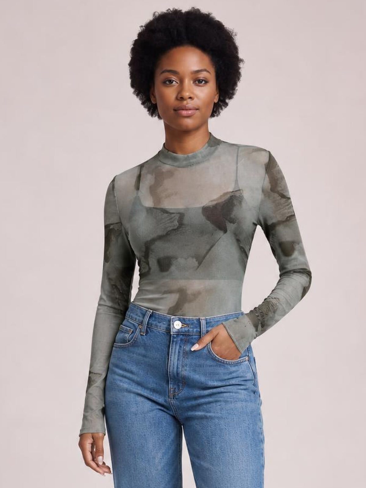 A model stands with one hand in her jeans pocket, wearing a fitted gray printed mesh T-shirt with a sheer mock neck, long sleeves, and a waist-length silhouette.