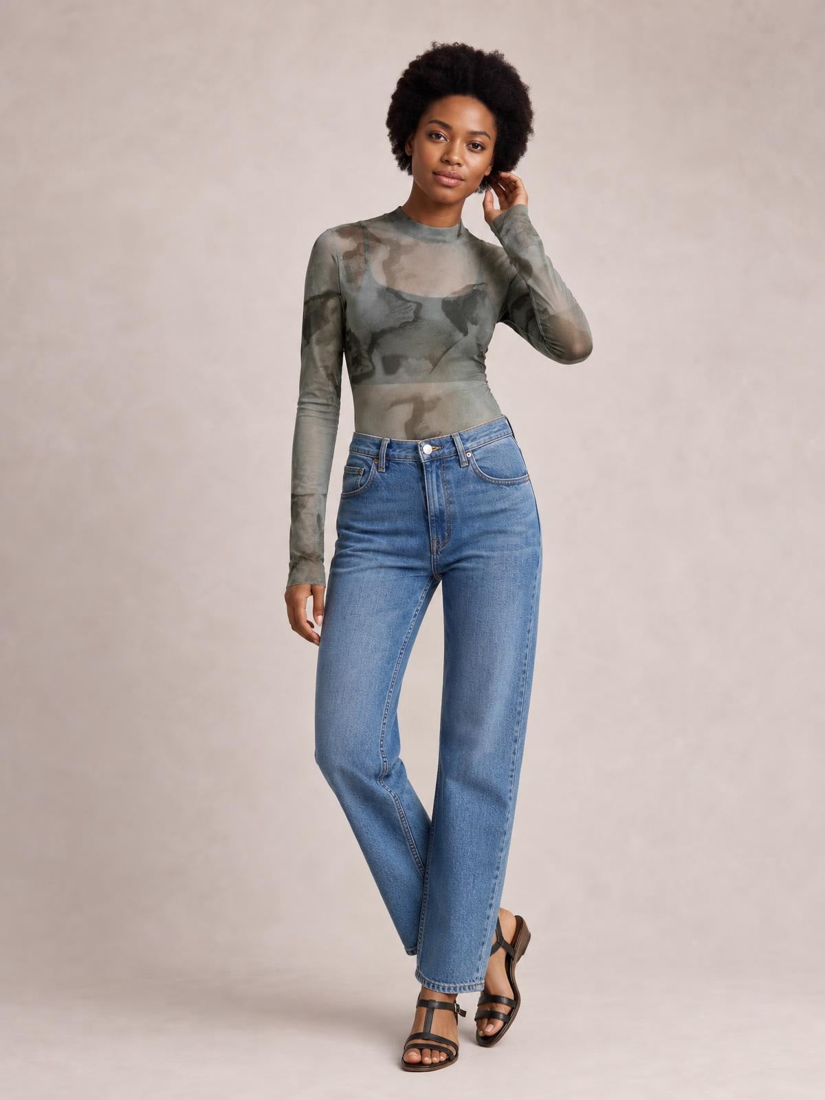 Model wearing a sheer printed mock neck long sleeve mesh T-shirt in a fitted silhouette, posing with one hand near her hair and crossed legs.