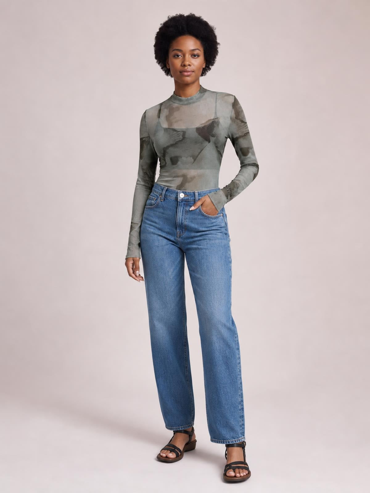 Model wears a gray-green printed sheer mock neck long sleeve mesh T-shirt with a fitted silhouette, standing with one hand in her jeans pocket.